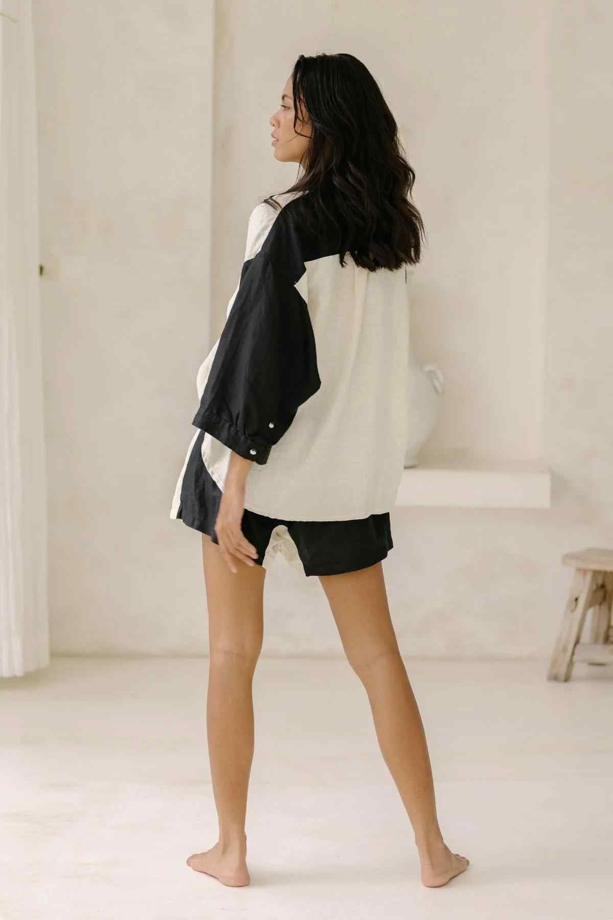 In a minimalist room with neutral-toned walls, simple decor, and a wooden stool on the right side, a woman with long dark hair stands barefoot. She is wearing the Yin Yang Nidra Button Down Set by Myrah Penaloza—a black and white oversized shirt made from sustainable fabric paired with black shorts.