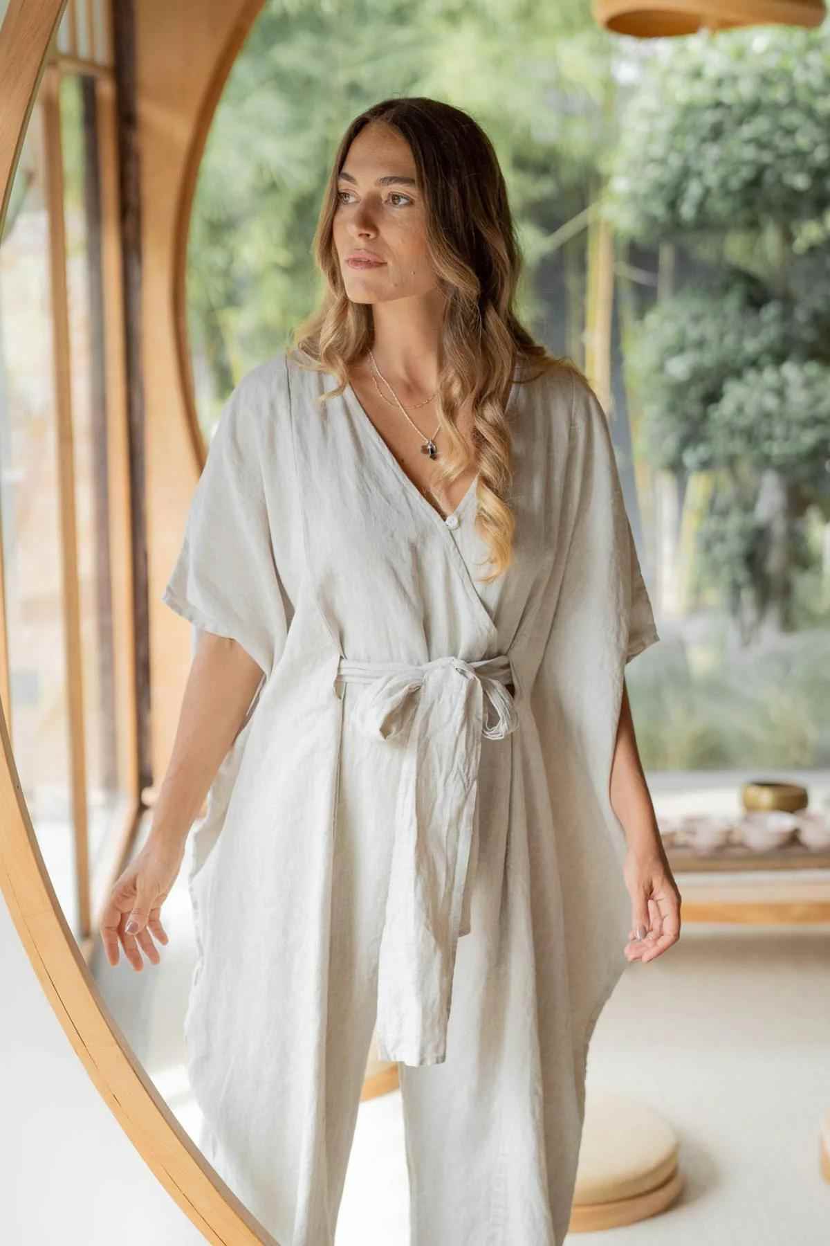 A woman with long, wavy hair wearing a light, flowing Linen River Playsuit by Myrah Penaloza stands by a large circular window. She gazes thoughtfully into the distance, with soft natural light illuminating her. A serene outdoor scene with greenery is visible through the window.