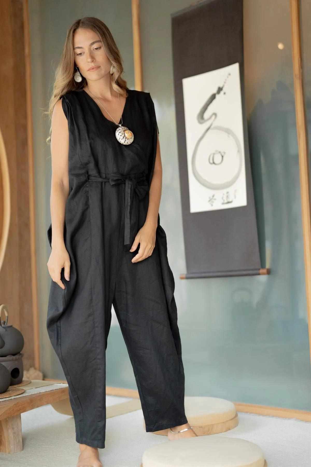 A woman with long hair stands barefoot indoors, showcasing the Myrah Penaloza French Linen Kuan Yin Playsuit with an adjustable waist tie and accessorized with a large shell necklace. She is next to minimalist decor, featuring a teapot-like object and a scroll adorned with an abstract design. The setting exudes a serene, modern aesthetic bathed in soft lighting.