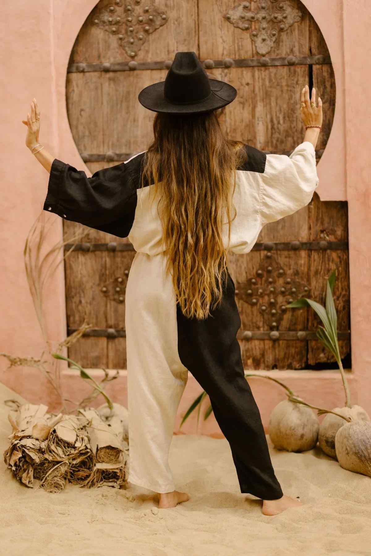 A person with long brown hair, wearing a black hat and the relaxed silhouette of the Yin Yang Nidra Button Down Set (Made To Order) by Myrah Penaloza, stands with their back to the camera, arms raised towards an ornate wooden door. Dried plants and coconuts are scattered on the sandy ground around them.