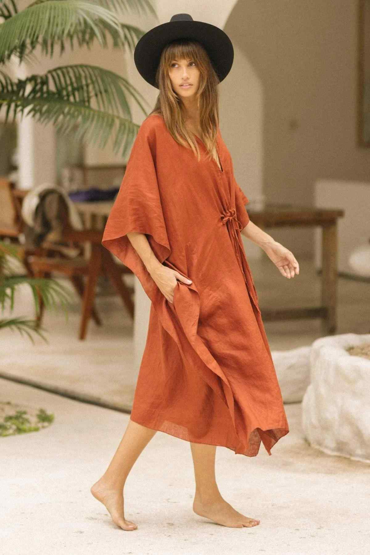 A woman with long hair, adorned in a wide-brimmed black hat and a flowing rust-colored Linen River Kaftan by Myrah Penaloza, walks barefoot on a light-colored floor. She is in a spacious, minimalist indoor setting featuring plants and wooden furniture.