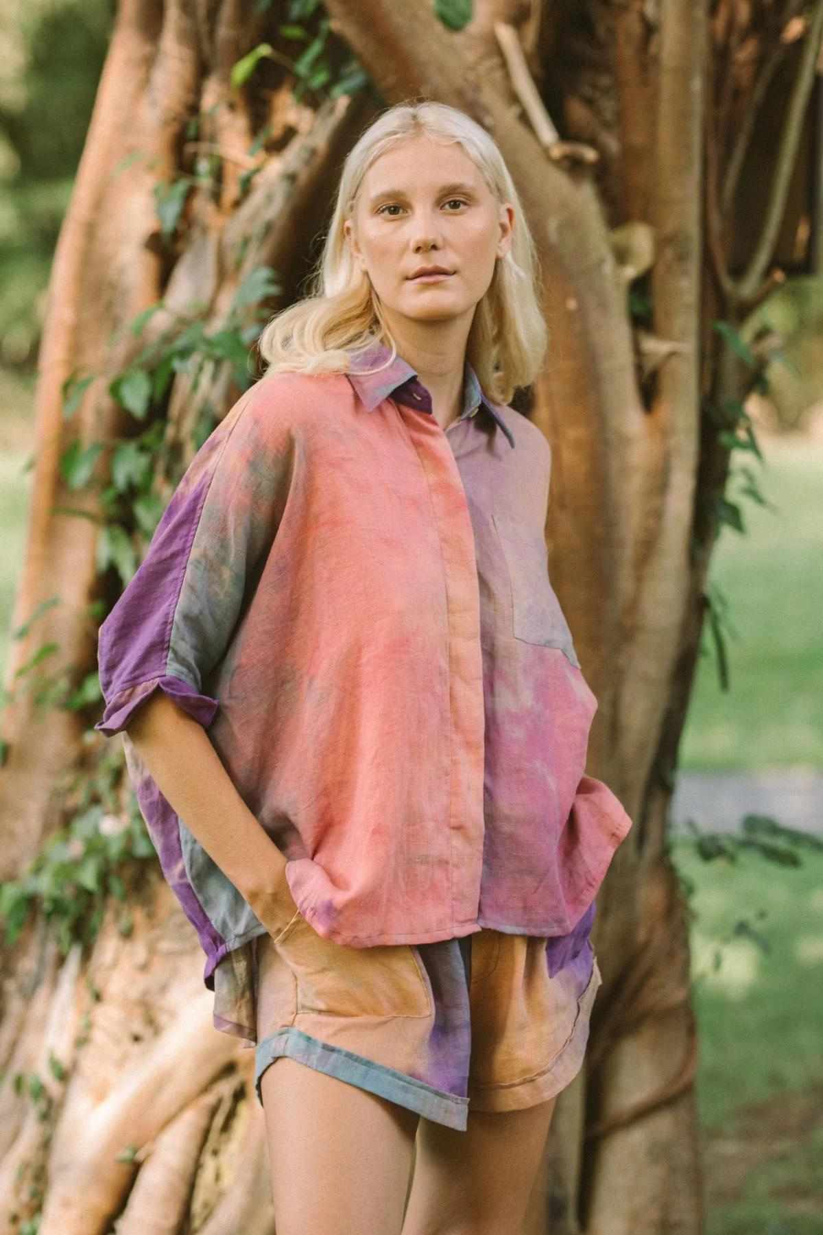 A person with light blonde hair stands outdoors in front of a tree, wearing the colorful, loose-fitting Rainbeau Linen Suka Set by Myrah Penaloza. The set consists of a tropical hand-dyed shirt with rolled-up sleeves and matching shorts. The backdrop is a natural setting with lush greenery, and the person has a neutral expression.