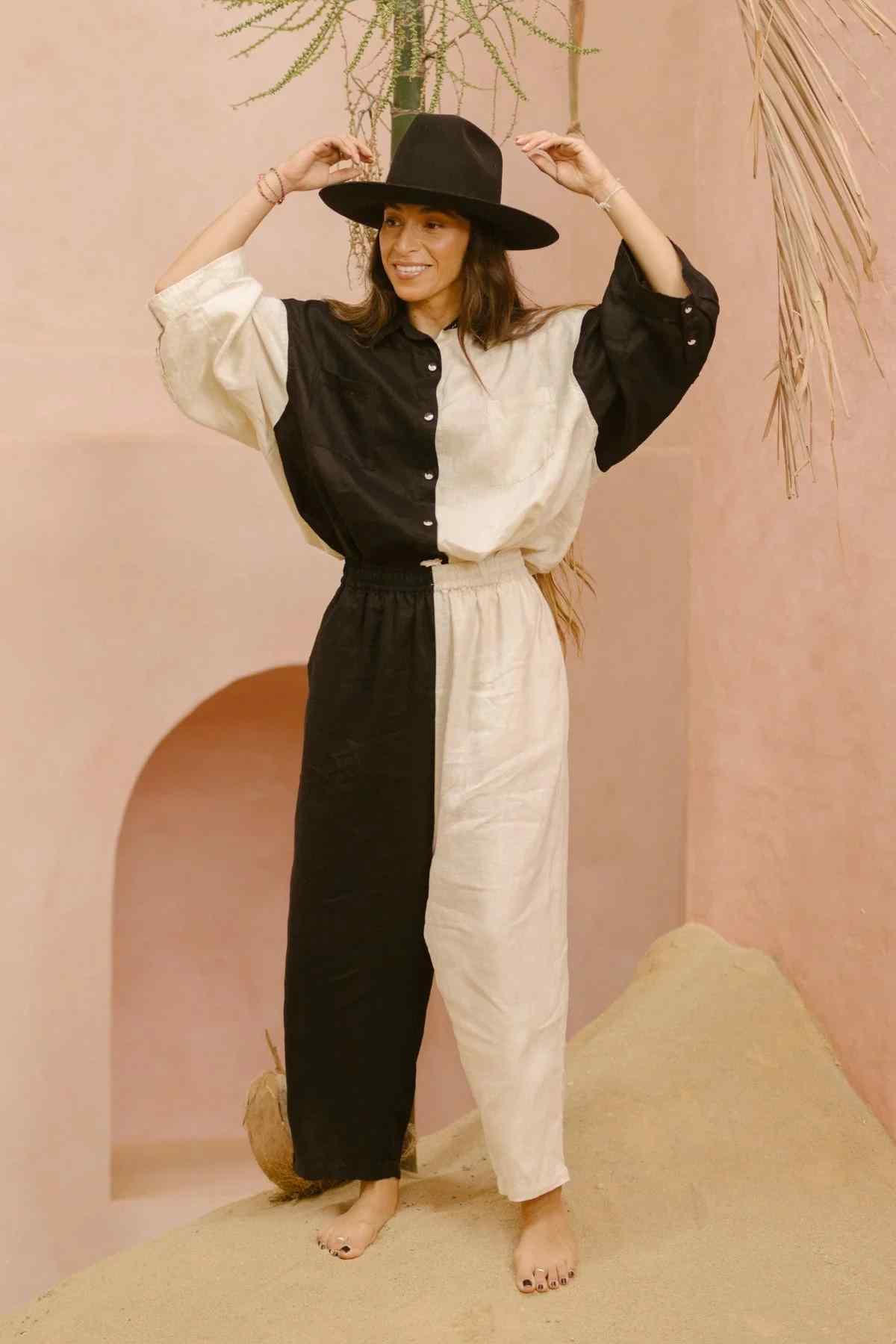 A person stands barefoot on a sandy surface inside a minimalist room with pale pink walls. They wear a wide-brimmed black hat and the Myrah Penaloza Yin Yang Nidra Button Down Set (Made To Order), featuring a relaxed silhouette in sustainable fabric with a half-black, half-white linen button-down. One hand touches the hat's brim while they smile and look away.