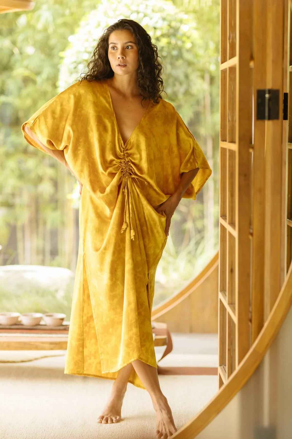 A woman stands indoors near a large, circular window, embodying a serene lifestyle. She wears the Silk Virgo Kaftan Botanically Dyed from Myrah Penaloza with a gathered front made of 100% silk in a flowing mustard yellow. Her curly hair is loose, complementing the green foliage and wooden, Japanese-inspired interior's open and airy feel.