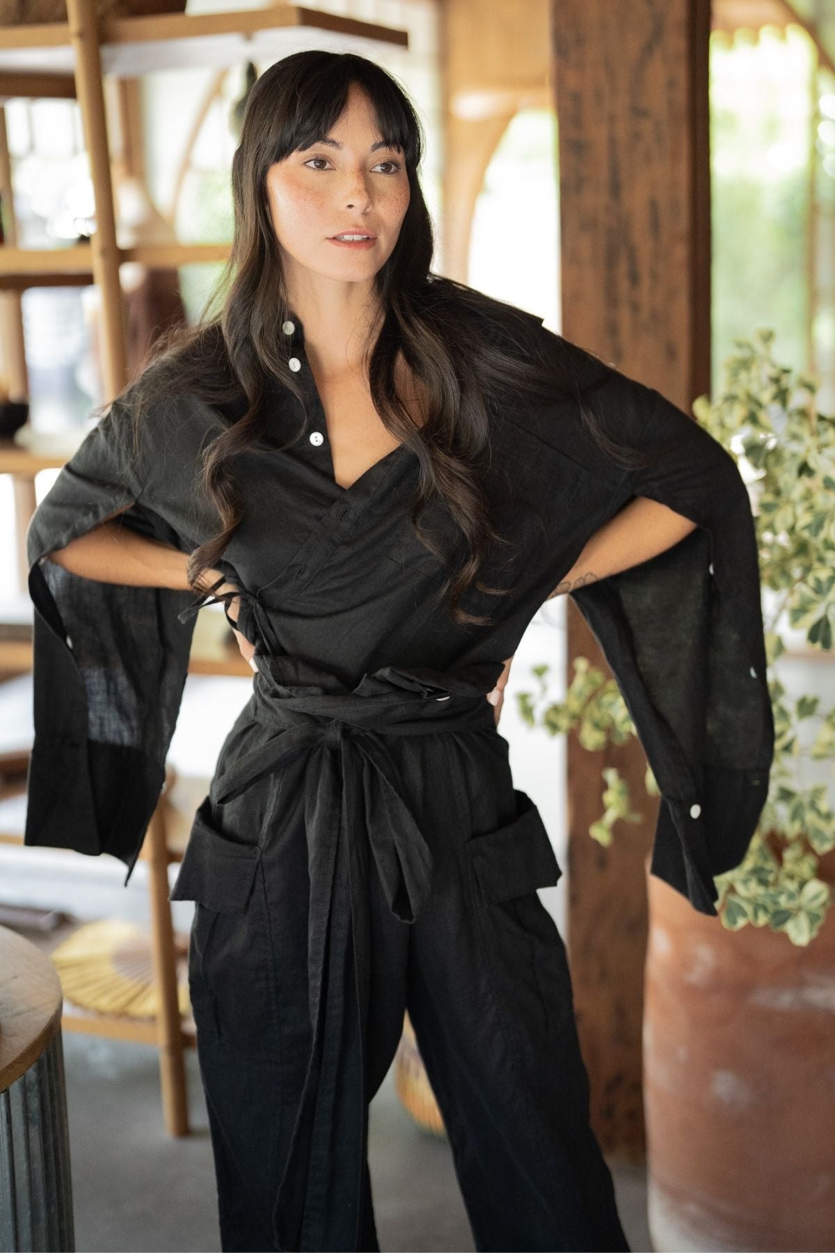 A woman with long, dark hair stands confidently with her hands on her hips, wearing the stylish black belted outfit with wide sleeves from the Sutra Linen Set by Myrah Penaloza. She is indoors, next to a wooden shelf adorned with potted plants, and gazes slightly to the side.