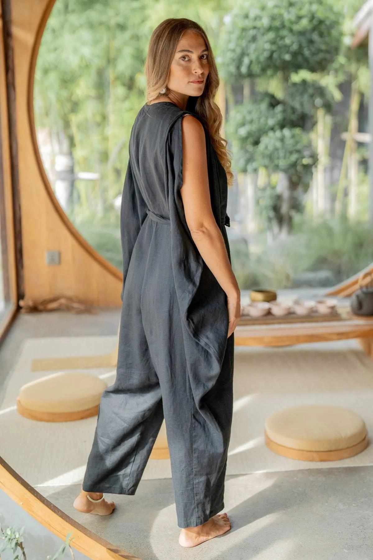 A woman with long hair stands barefoot in a serene room with a circular window, revealing a lush green garden. She wears the French Linen Kuan Yin Playsuit by Myrah Penaloza, which features an adjustable waist tie, and gazes back over her shoulder. The room is adorned with round cushions and minimalist wooden decor.
