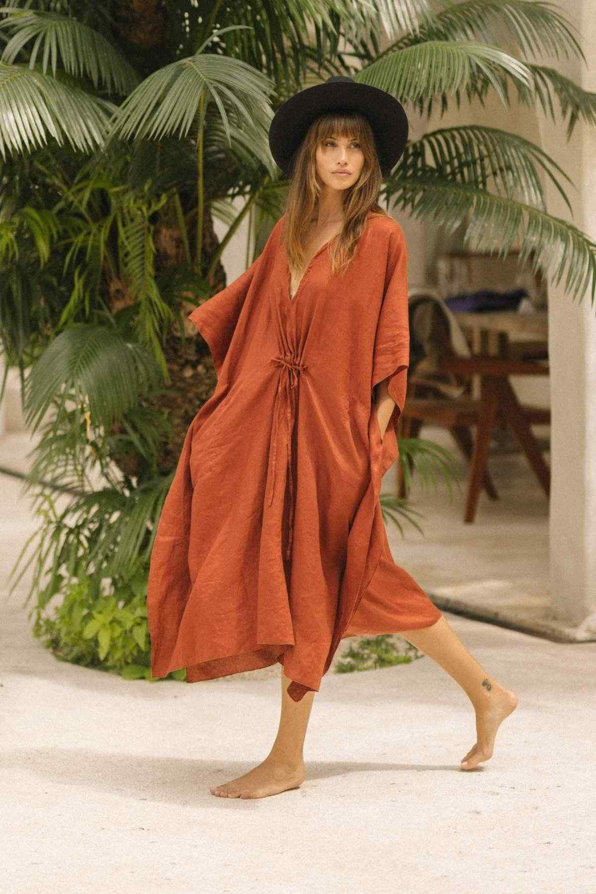A woman in a wide-brimmed black hat and a loose, rust-colored Linen River Kaftan by Myrah Penaloza walks barefoot on a sandy path. She is surrounded by lush green tropical plants, with a wooden chair visible in the background.
