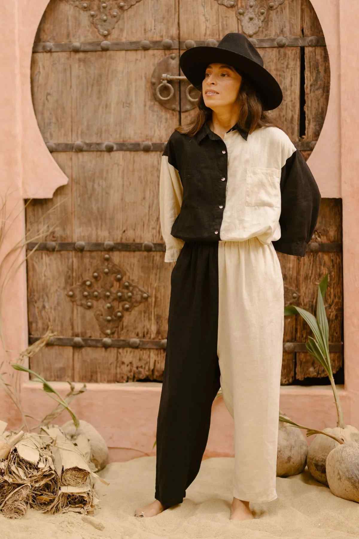 A person in the Yin Yang Nidra Button Down Set from Myrah Penaloza, featuring a black and white color-blocked design, stands barefoot on sand in front of a rustic wooden door adorned with decorative metal accents. Their relaxed silhouette harmonizes perfectly with the scattered dried palm leaves and driftwood in the background, all while sporting a wide-brimmed black hat.
