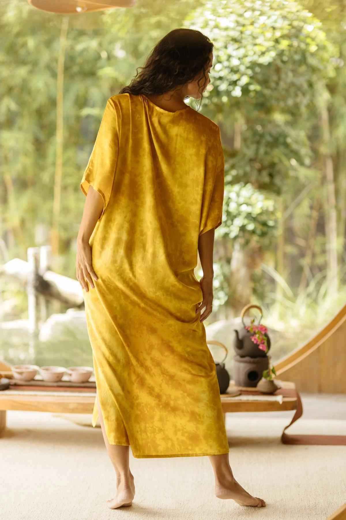 A person stands barefoot on a wooden floor, adorned in a loose, Silk Virgo Kaftan Botanically Dyed in golden-yellow from Myrah Penaloza. They face away with their head turned slightly to the side. The background reveals a lush garden viewed through large windows, with a tea set arranged on a table nearby, evoking an elegant lifestyle.