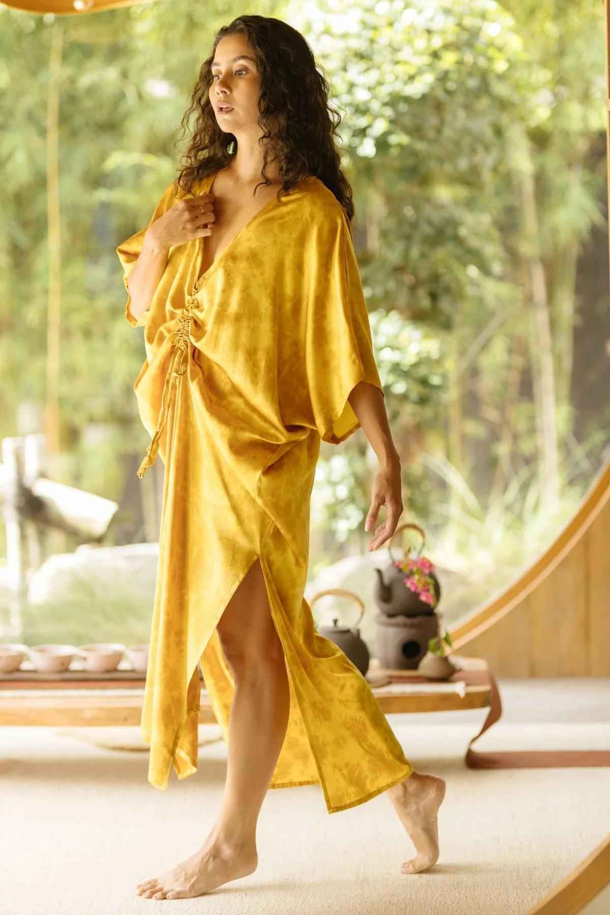 A serene woman stands barefoot indoors, showcasing the Silk Virgo Kaftan Botanically Dyed by Myrah Penaloza. The golden yellow kaftan, made from 100% silk, features a loose-fitting design with a ruched front and high slit. Her long wavy hair adds to her elegant appearance as she embodies relaxed lifestyle elegance. In the background, a teapot rests on a low table near a large circular wooden window that frames lush greenery outside.