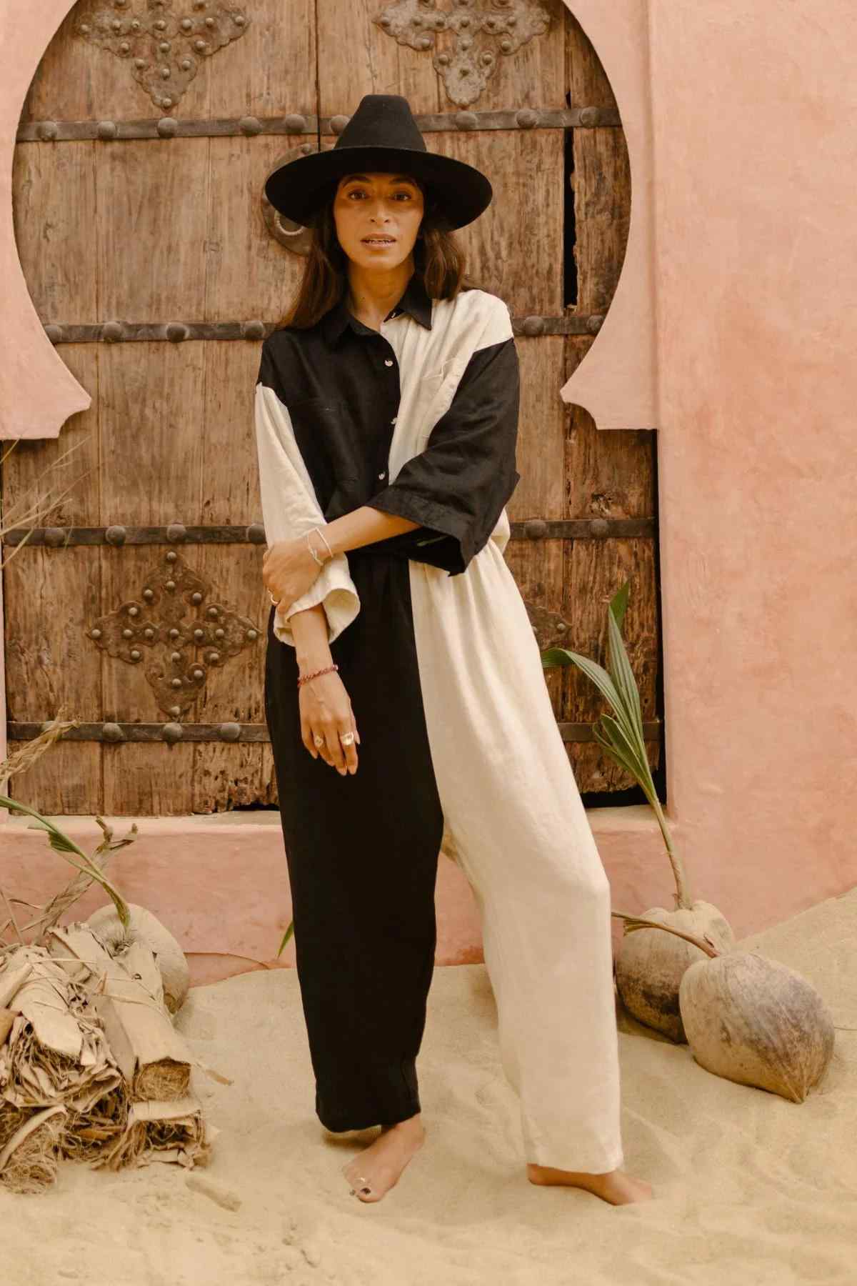 A woman stands barefoot on sandy ground in front of an arched wooden door. She wears a black wide-brimmed hat and the Yin Yang Nidra Button Down Set from Myrah Penaloza, featuring a split black and white color scheme with a relaxed silhouette. Surrounding her are plants and coconuts, contrasting against the pink-toned walls in the background.