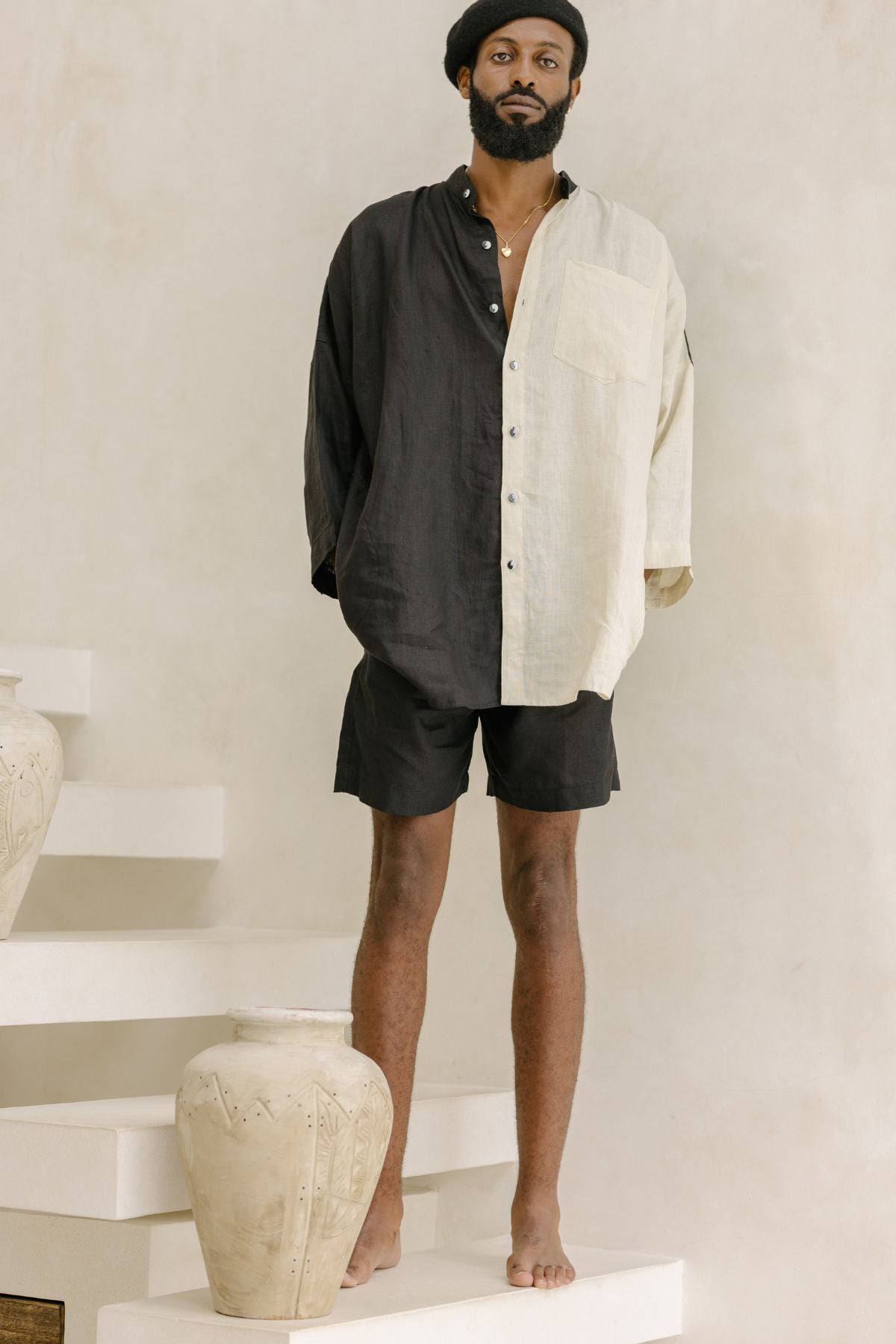 A man stands on white steps next to large pottery, wearing a half-black, half-white "Camisa del Novio" linen top from Myrah Penaloza and black shorts. He is barefoot and wearing a black beret against a neutral and minimalistic background.