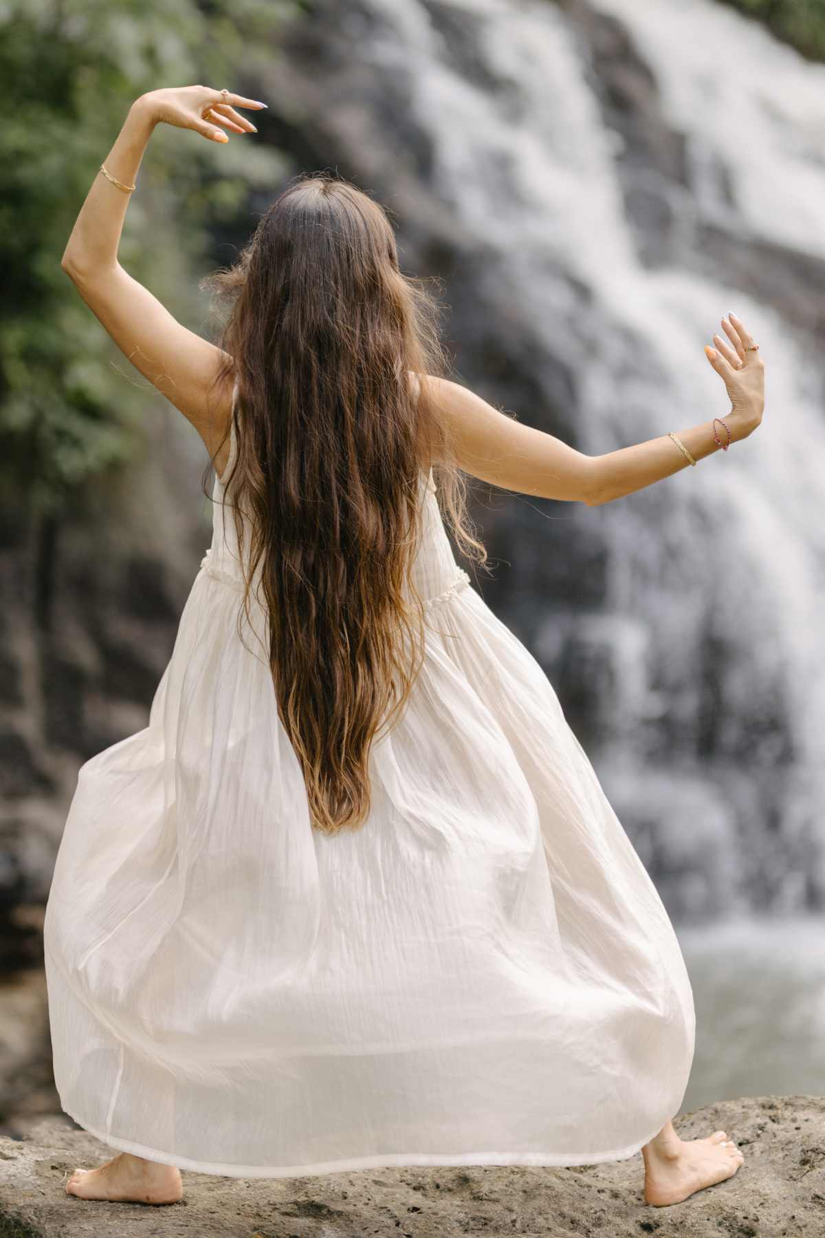 A woman with long, wavy hair is dancing barefoot on a rock in front of a waterfall, wearing the flowing Dolly Night Gown by Myrah Penaloza. Her arms are gracefully raised, creating a fluid and serene scene with nature as the backdrop.