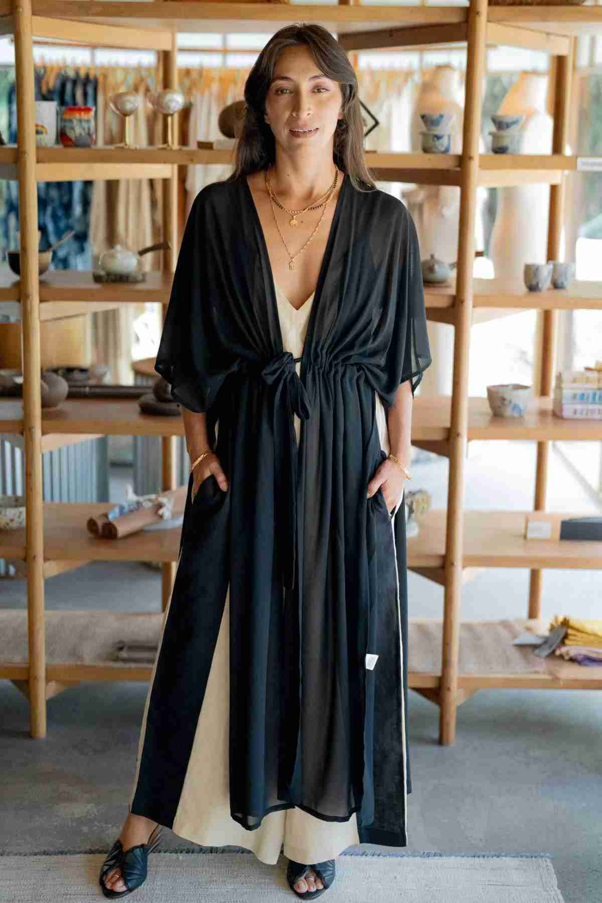 A woman with long dark hair stands in a store, radiating elegance as she wears the Madelin Sheer Chiffon Long Cover by Myrah Penaloza over a white dress. With her hands in the cover's pockets and adorned with multiple necklaces, she exudes sheer brilliance. Shelves filled with various items are visible in the background.