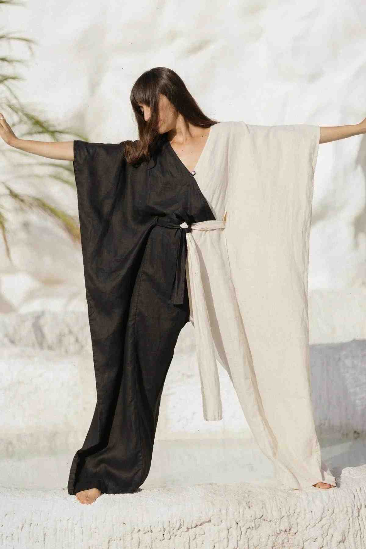 A person with long hair is posing with their arms extended, wearing the French Linen Yin Yang River Playsuit by Myrah Penaloza. The outfit features a two-toned kimono-style design split into black on one side and white on the other. The background consists of a light, natural setting with a hint of greenery, reflecting a sense of eco-consciousness.