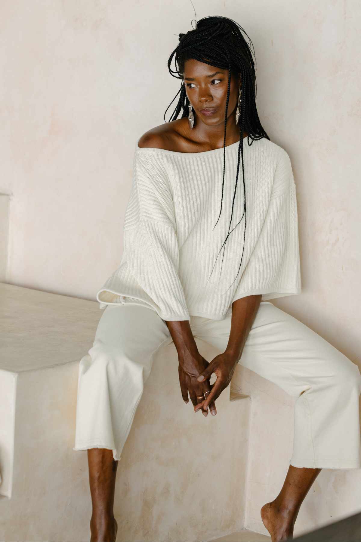 A person with long braided hair is seated on a light-colored step against a neutral background. They are wearing the luxurious Spirit Top Sweater Knit from Myrah Penaloza, an oversized, off-shoulder white sweater made of premium cotton fabric, paired with matching wide-legged trousers. The person looks downwards, with their hands resting on their lap.