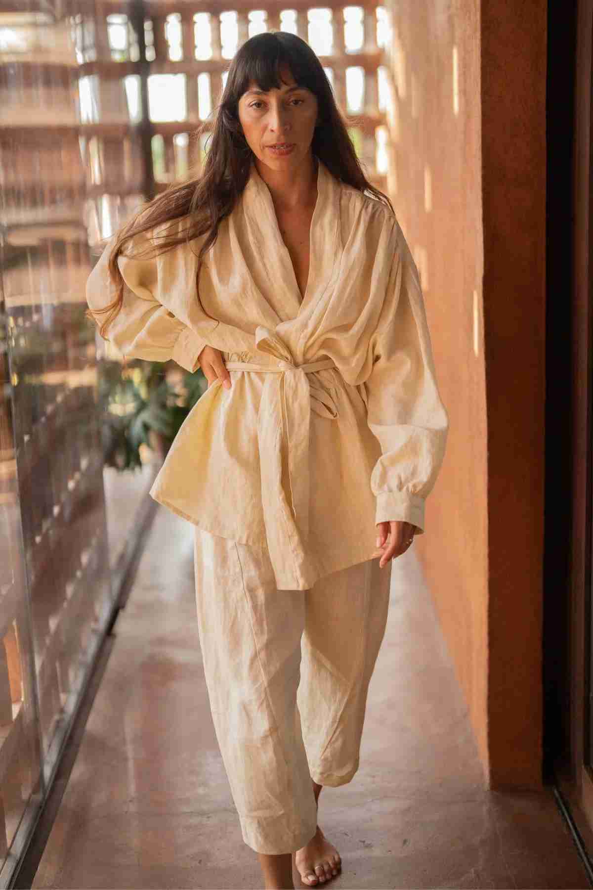 A woman with long brown hair, walking barefoot indoors, is wearing a cream-colored, belted, loose-fitting top and matching pants from the Linen Jasmine Set by Myrah Penaloza. The kimono-inspired design and serene ambiance of wooden structures and glass panels create a warm, natural feeling. She has a confident expression.