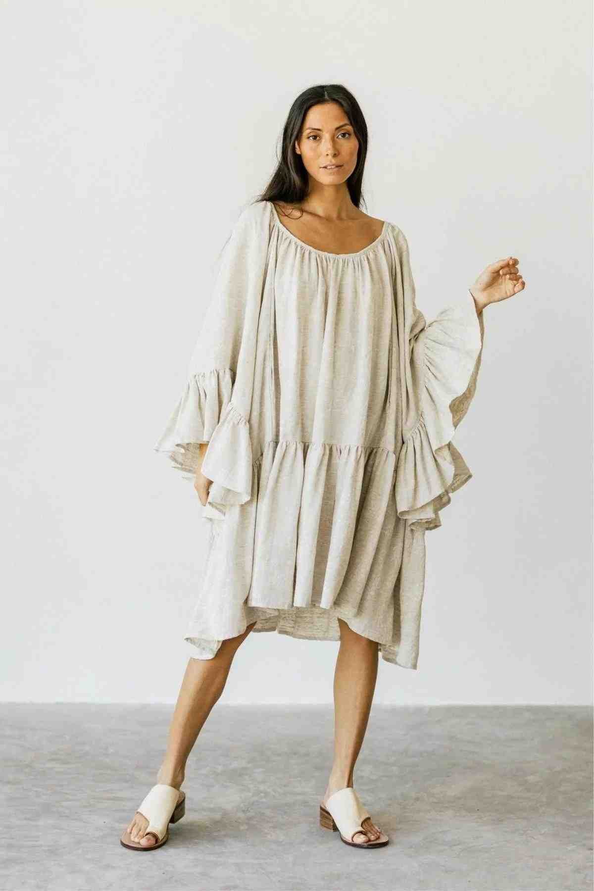 A person poses gracefully against a minimalistic backdrop, wearing the Linen Brida Gown Short by Myrah Penaloza. Crafted from linen with wide sleeves and ruffled accents, this neutral-colored dress captures the romantic essence of spring/summer style. Complementing the gown are open-toed sandals that complete the airy ensemble.