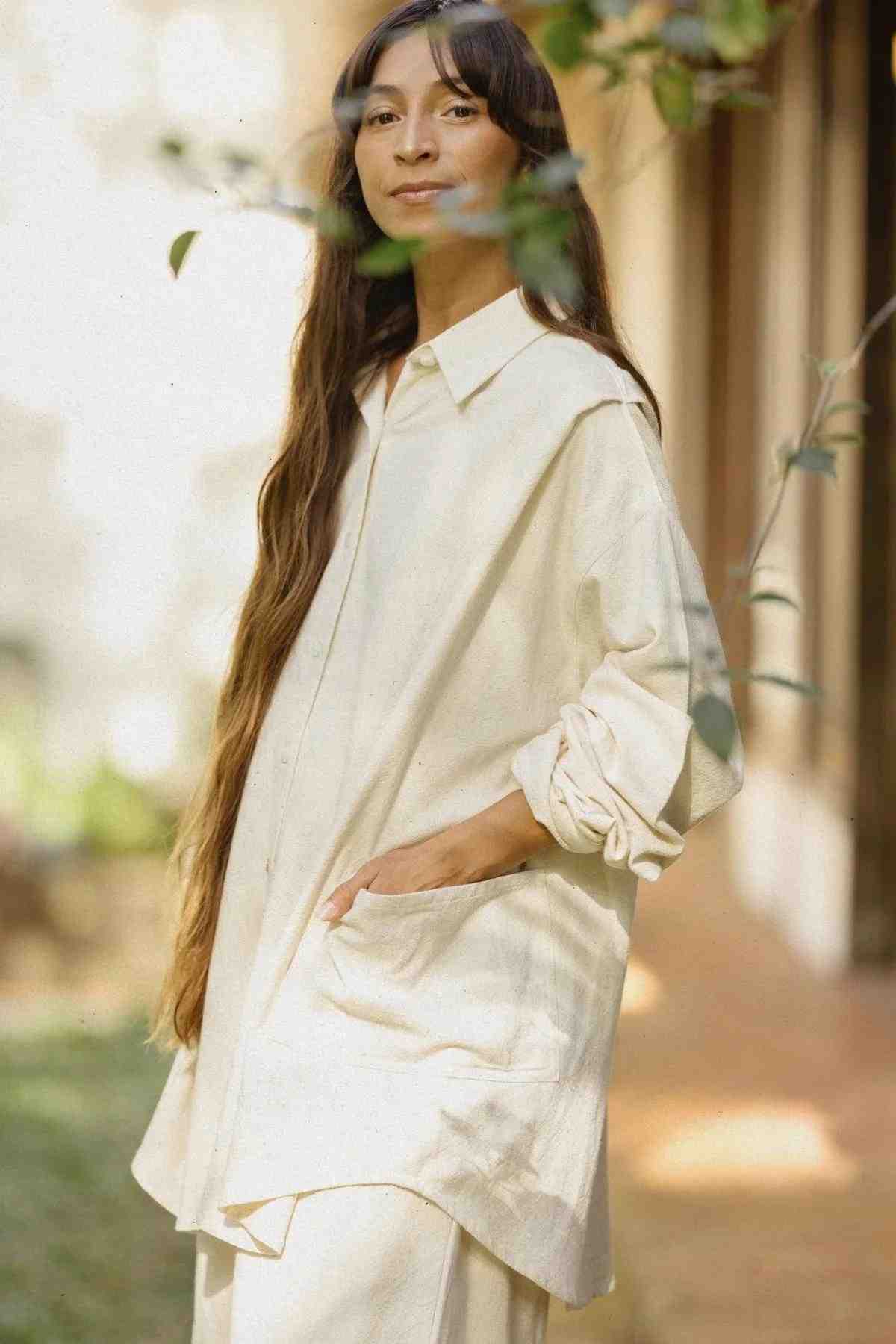 A person with long, dark hair stands outdoors wearing a light-colored, oversized shirt with rolled-up sleeves and matching pants. They have a serene expression and an eclectic piece—a Myrah Penaloza Tender Jacket in Deep Royal Blue (100% Linen)—draped over one shoulder. The background appears to be a sunlit garden or outdoor area with greenery.