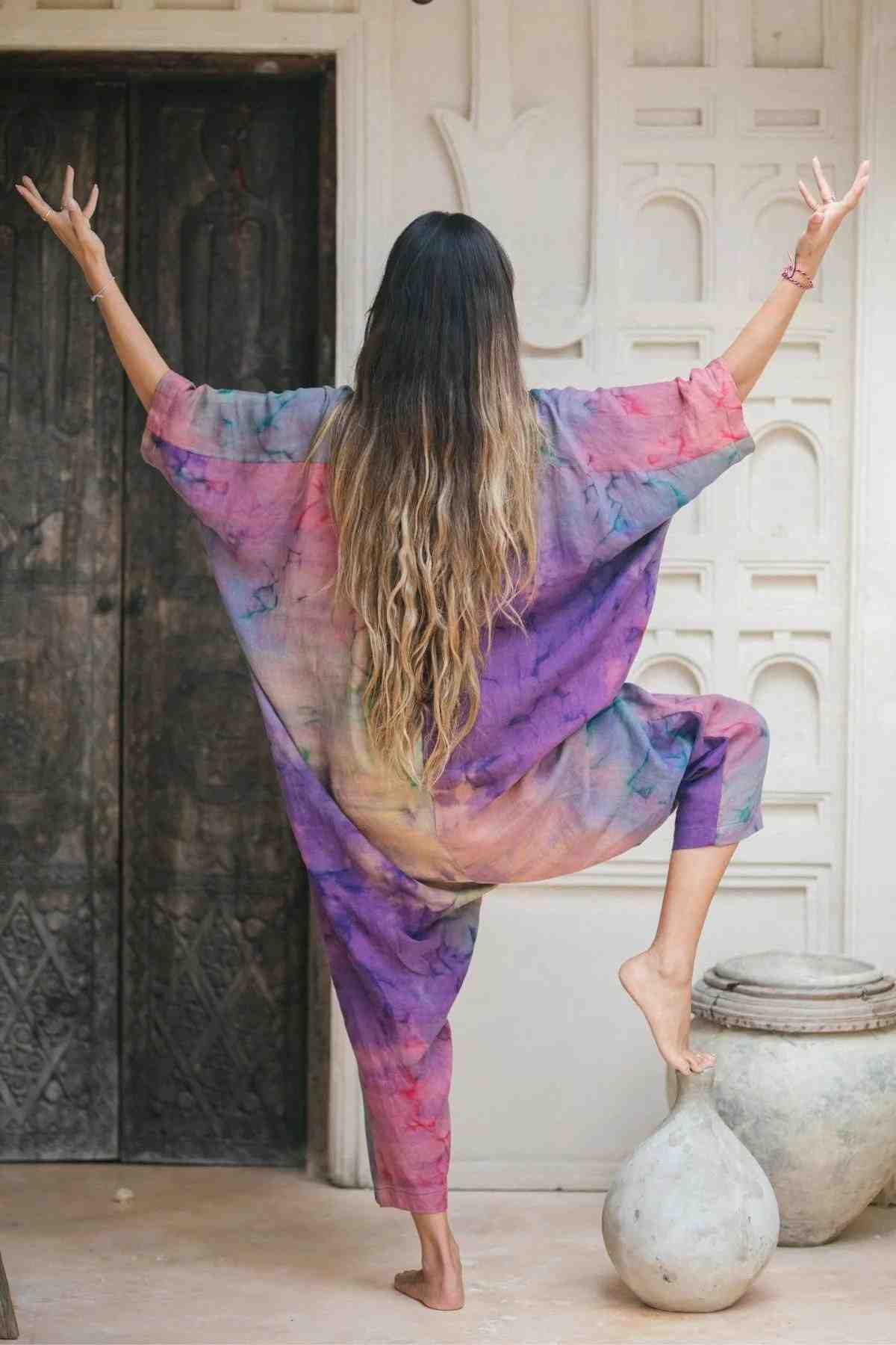 A person with long hair stands on one leg in a yoga pose, raising their arms in the air. They are wearing the colorful, loose-fitting Sat Torri Rainbeau Playsuit by Myrah Penaloza, featuring shades of pink, purple, and blue reminiscent of retro 1980's design. The background showcases an ornate door and white walls with decorative patterns.