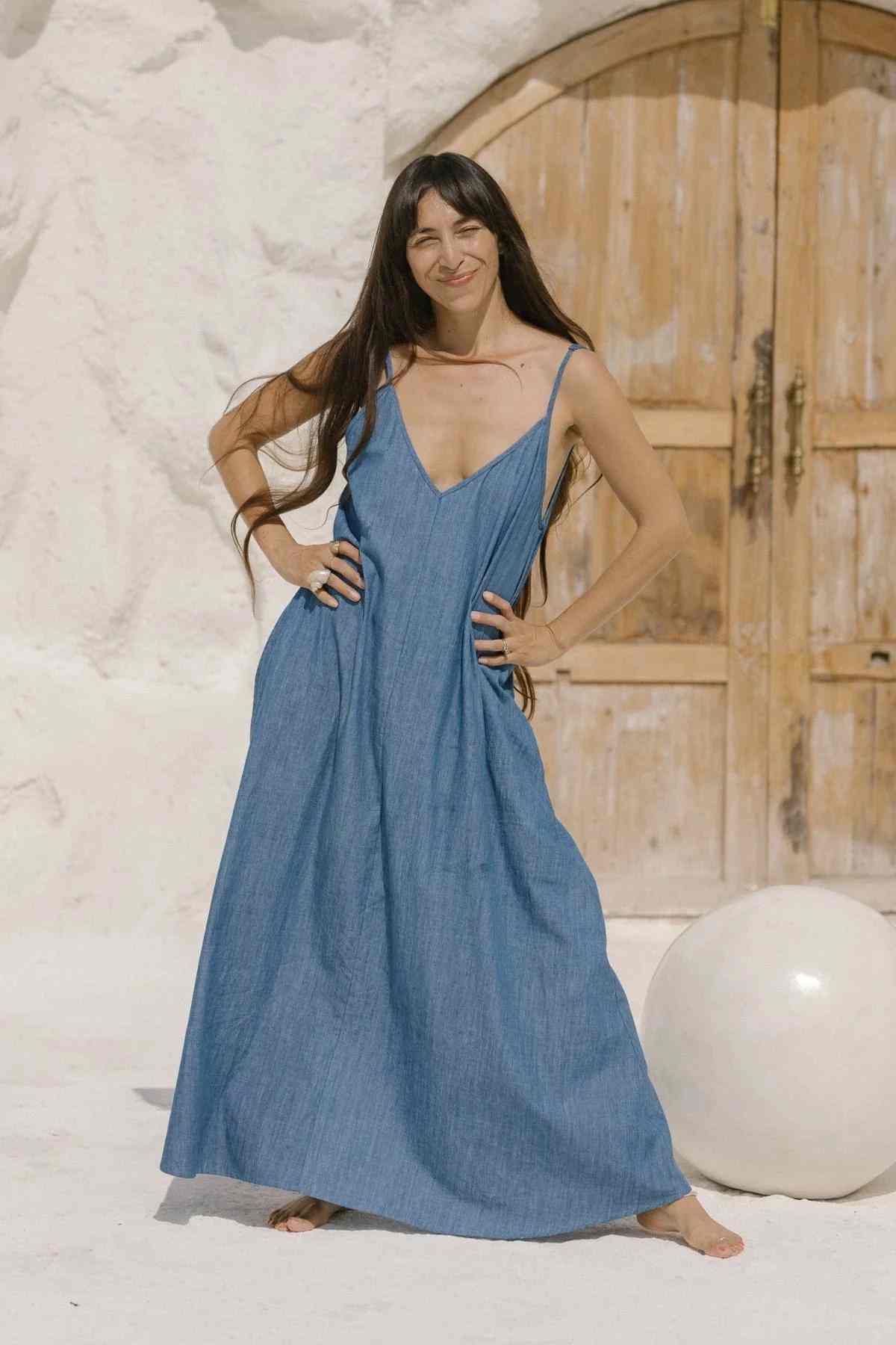 A woman with long dark hair stands barefoot in front of a rustic wooden door. She is wearing an exquisite Kundalini Denim Dress by Myrah Penaloza, made of stonewashed linen and sleeveless, with one hand on her hip. The background is a light-colored, textured wall, and there is a white spherical object near her feet.