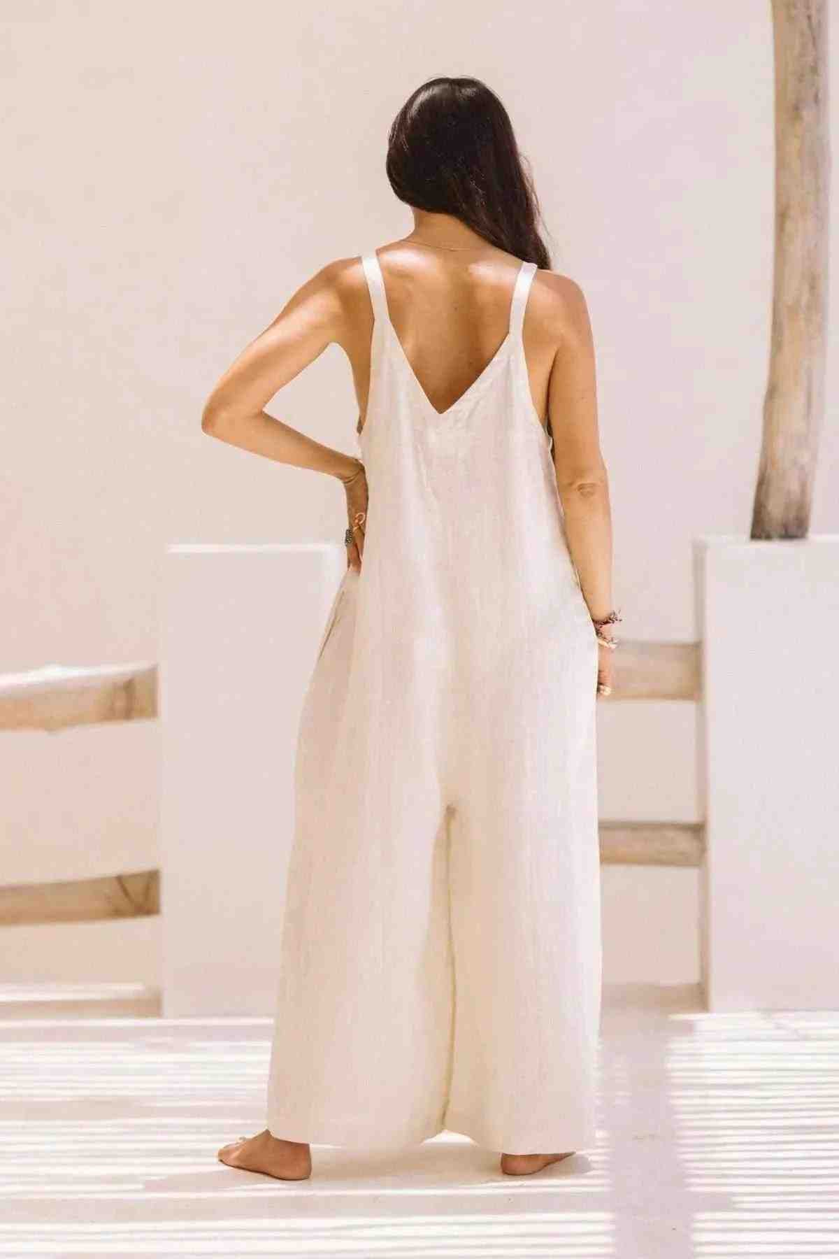 A person with long hair is standing with their back to the camera, wearing the Kundalini Linen Playsuit by Myrah Penaloza. Made of luxuriously soft stonewashed linen, this loose, white sleeveless jumpsuit is showcased in a sunlit, minimalistic setting filled with light shadows and wooden elements in the background.
