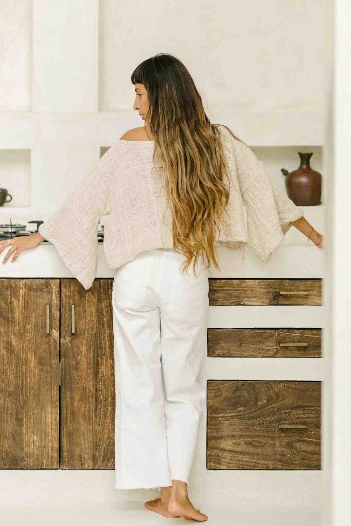 A person with long, wavy hair stands barefoot in a kitchen, facing away from the camera. They are wearing an oversized Tumanggal Cotton Spirit Top by Myrah Penaloza, made of durable fabric that exposes one shoulder and white pants. The kitchen features wooden cabinets and a white countertop oven.