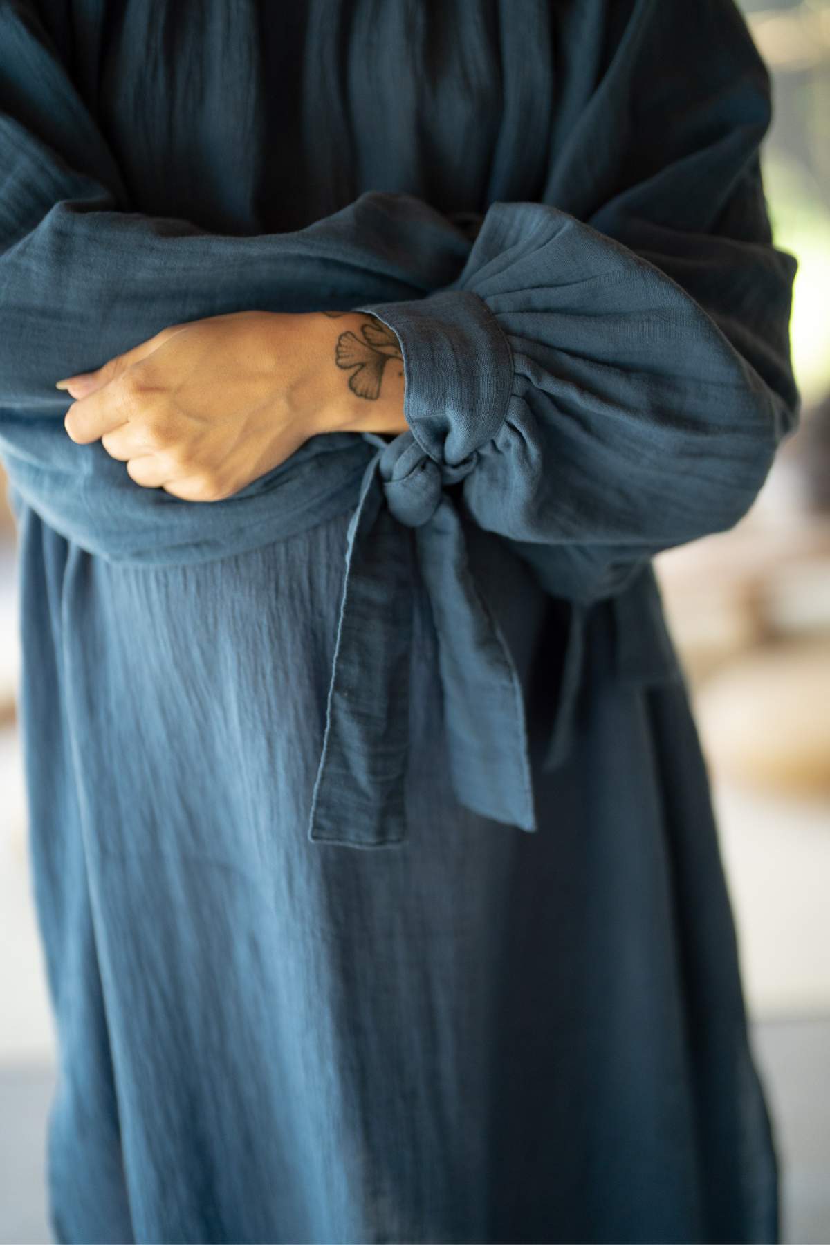 A person wearing the Longing Gown by Myrah Penaloza, a reversible A-line dress with long sleeves, stands with arms crossed. One sleeve is stylishly knotted, revealing a clover tattoo on the left wrist that subtly alludes to Virgo details. The background is artistically blurred.