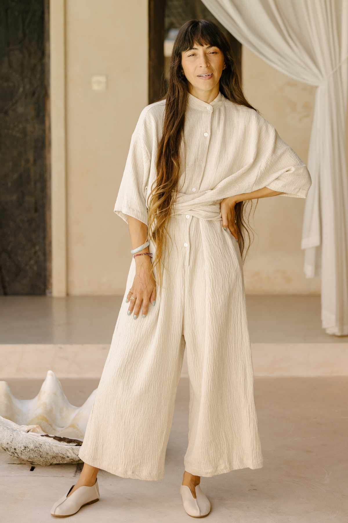 Dressed in the Pine Playsuit by Myrah Penaloza, a person stands indoors with long, wavy hair and exudes effortless style. One hand rests on their hip near a decorative shell, while draped curtains provide an elegant backdrop for this versatile wardrobe staple.