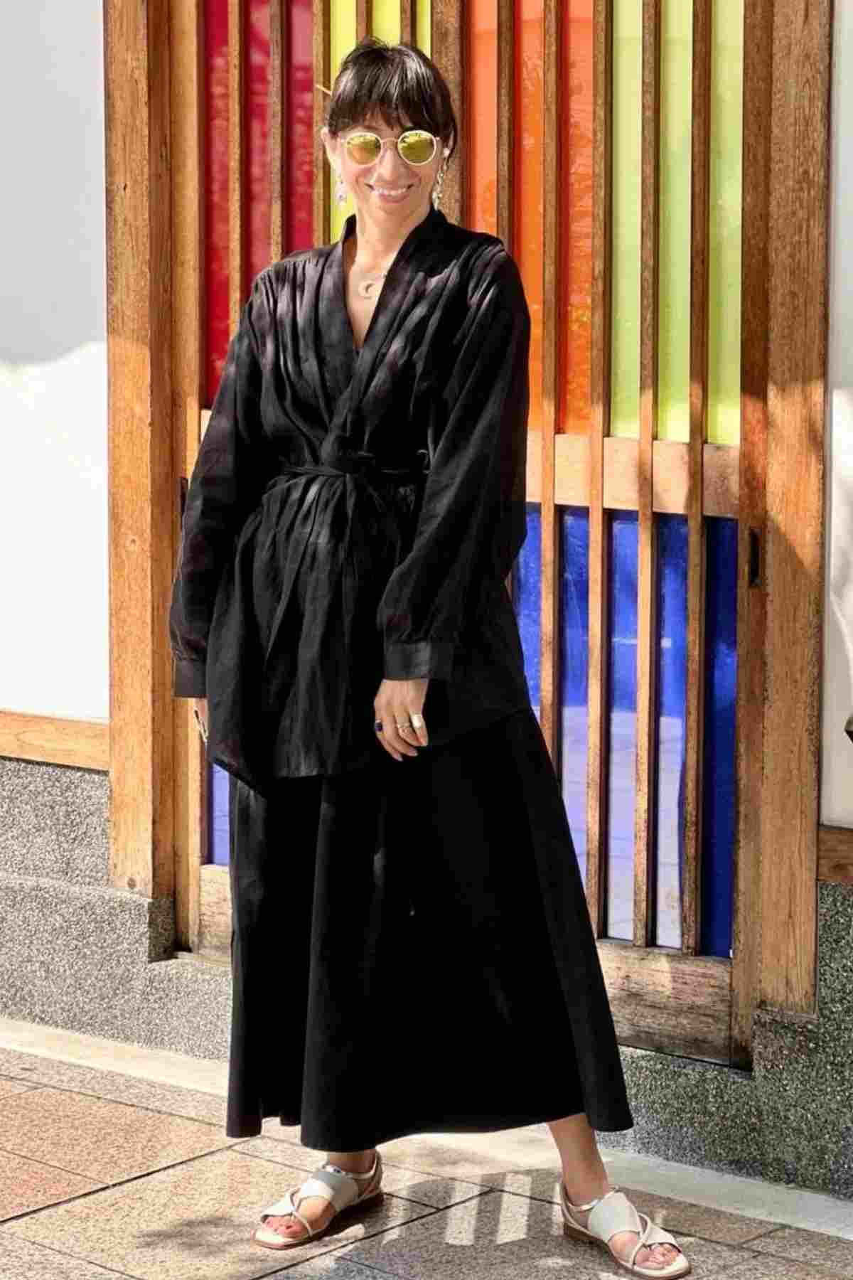 A person wearing oversized sunglasses and a black belted outfit stands in front of a colorful wooden panel with vertical stripes. They smile confidently on the stone pavement, their white sandals highlighted by Pantalon Linen Pants (Wide Legged) from Myrah Penaloza, which contrast the dark attire.
