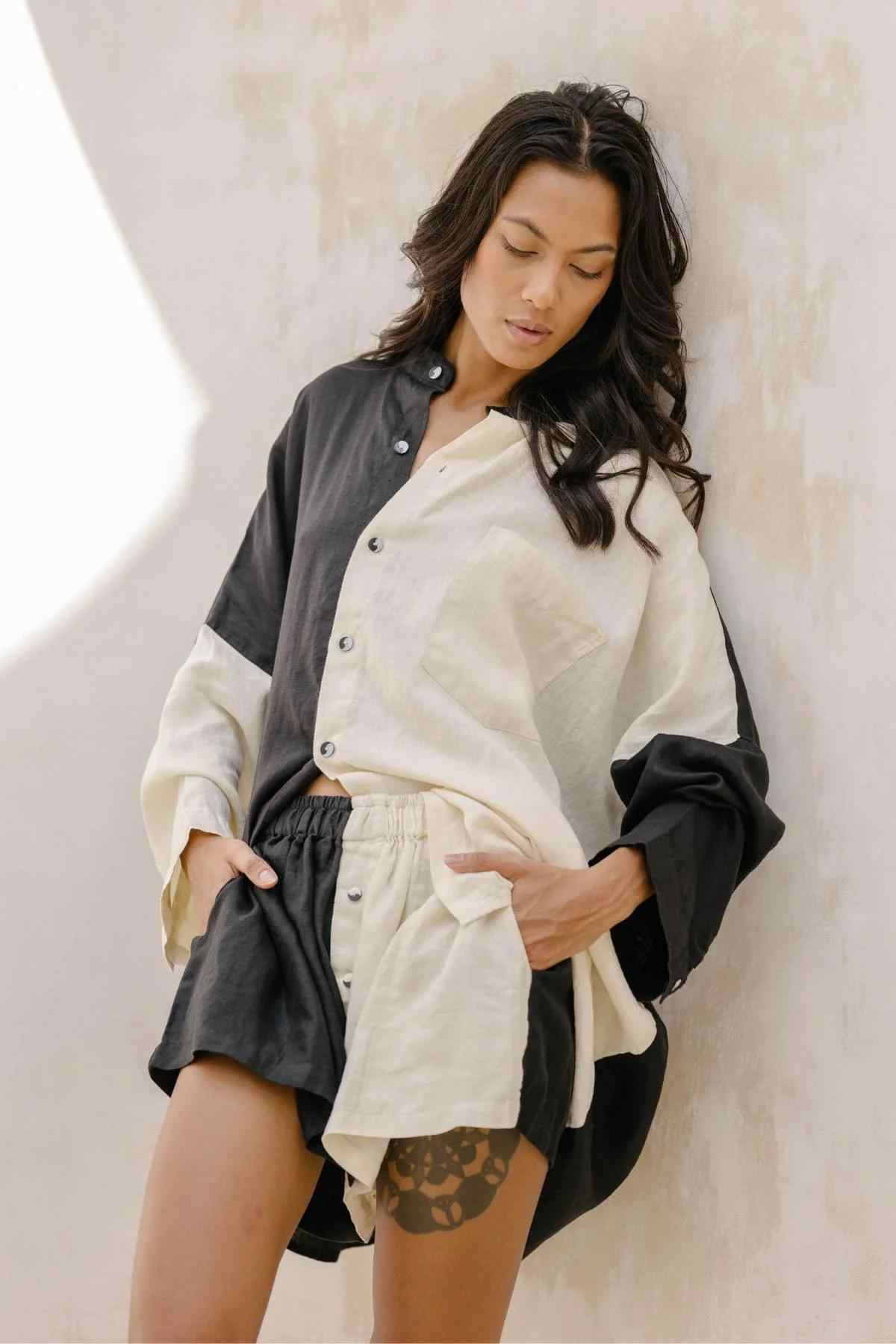 A woman with long dark hair leans against a textured off-white wall, elegantly dressed in the Unisex Yin Yang Linen Camisa Del Novio Set by Myrah Penaloza. Her color-blocked outfit consists of a buttoned shirt and matching shorts, primarily in black and white. Crafted from indigo dyed fabric, her handmade clothing catches the subtle sunlight, creating a soft glow on the wall.