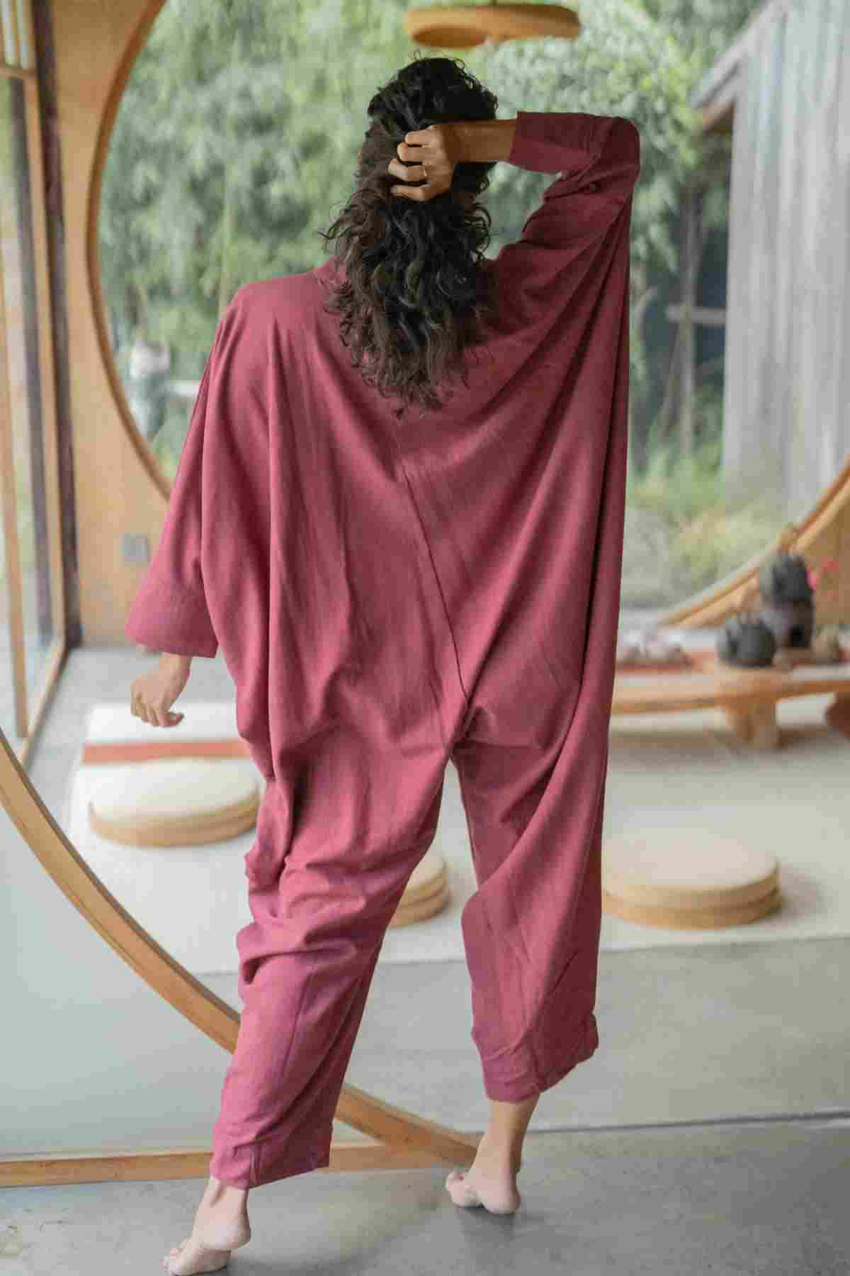 A person with curly hair stands indoors, facing away, with one hand on their head. They are wearing a loose, flowing red outfit from the Miracle Playsuit 2.0 (Karama Collection) by Myrah Penaloza. The room features round windows, wooden floors, and minimalistic decor, creating a serene atmosphere with greenery visible outside.