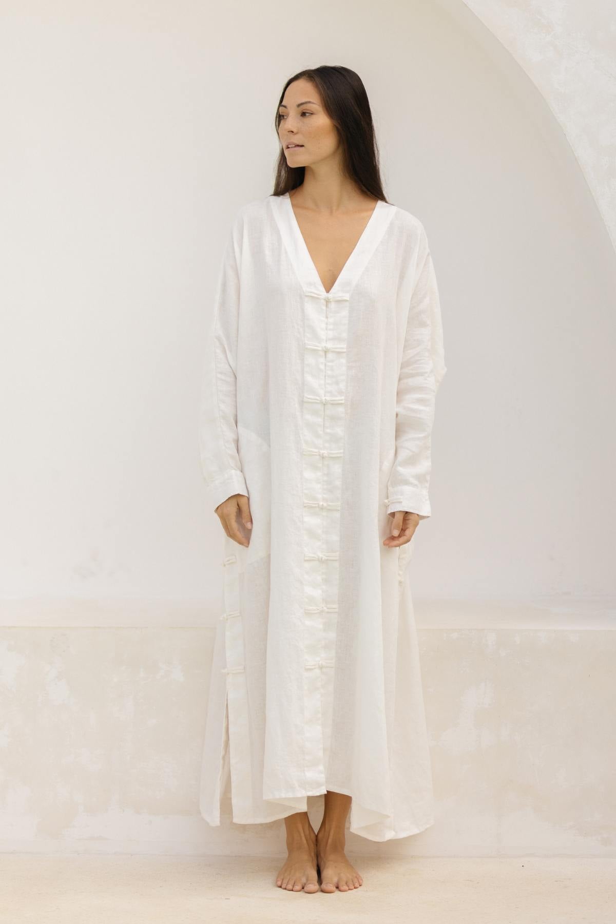A woman stands barefoot against a plain backdrop, wearing the Ancient Chajin Jacket by Myrah Penaloza. The 100% linen jacket is long with a V-neck design, long sleeves, and front pockets. Japanese-inspired buttons add an elegant touch to the garment. She gazes to her right with a serene expression as her straight, dark hair falls naturally over her shoulders.