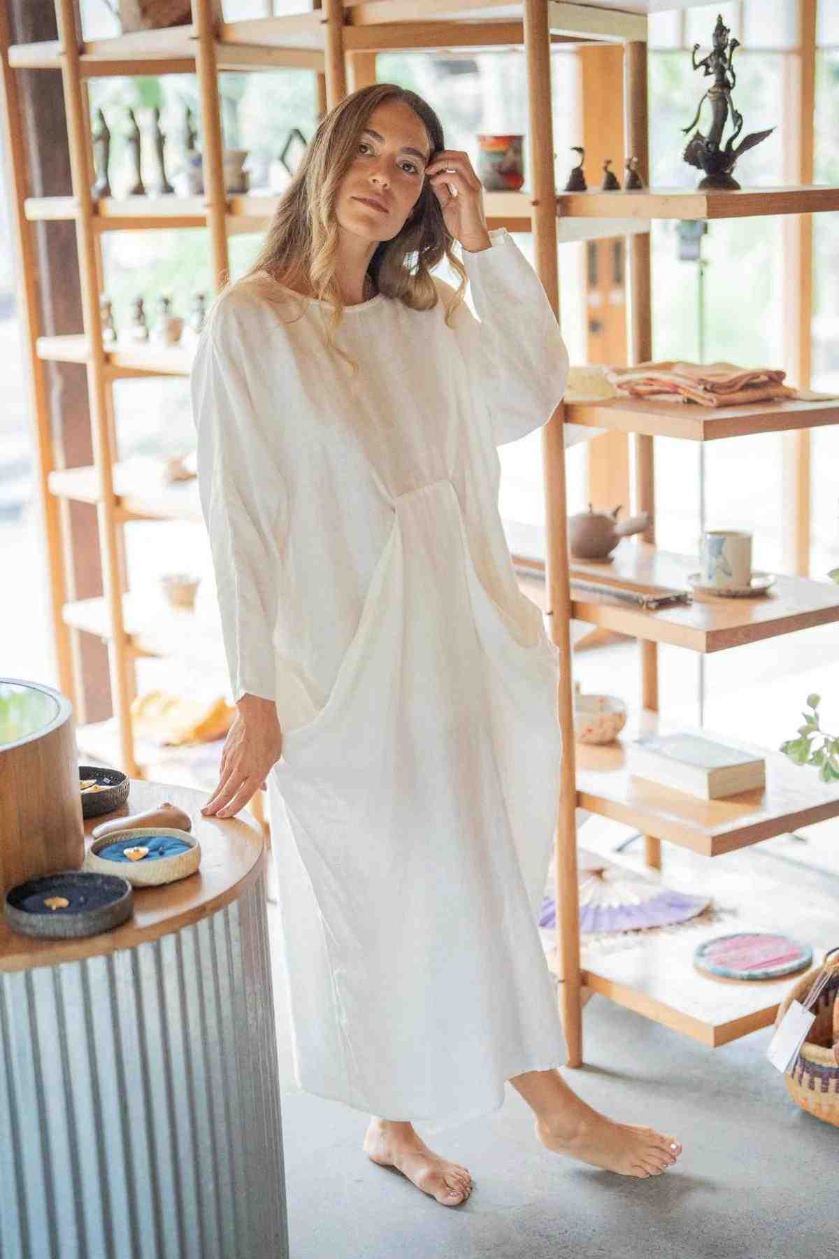 A woman wearing a Myrah Penaloza Mountain Magic Linen Gown stands barefoot in a stylishly decorated room with wooden shelves. She has long hair and is gently touching her head with one hand while looking at the camera. Various decorative items are displayed on the shelves behind her, adding charm to the scene.