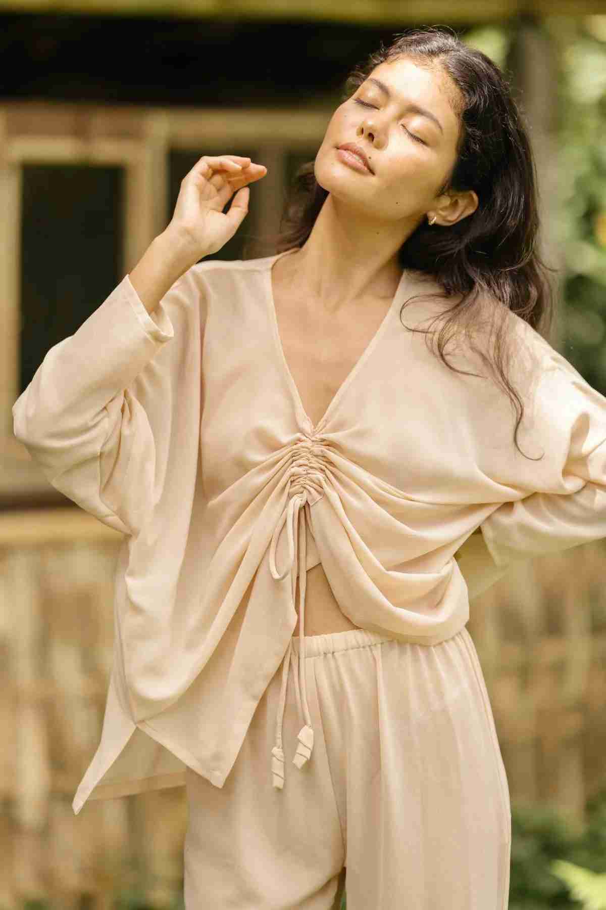 A woman with long, wavy hair is standing outdoors with closed eyes, wearing the Chiffon Silk Sheer Virgo Rising Set by Myrah Penaloza. She has a serene expression and one hand raised slightly as she enjoys the natural sunlight. The background features greenery and a rustic building.