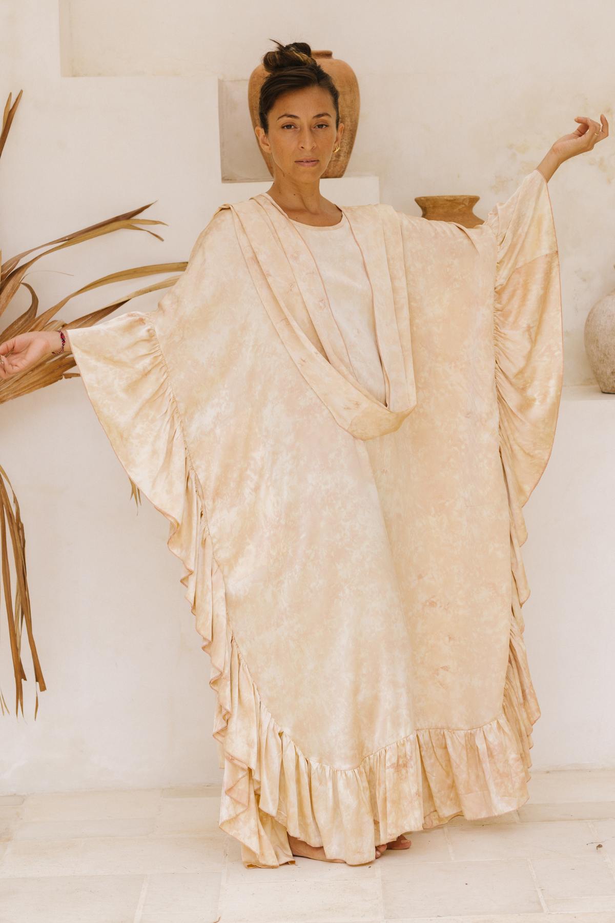 A person stands indoors, wearing the Myrah Penaloza Perla Silk Gown Made To Order in a flowing, light beige fabric with ruffled edges that evoke a Florence and The Machine aesthetic. Their hair is tied up, and their arms are slightly raised to the sides. The minimalist decor in neutral tones features pottery and tall dried palm fronds, creating an ethereal ambiance.