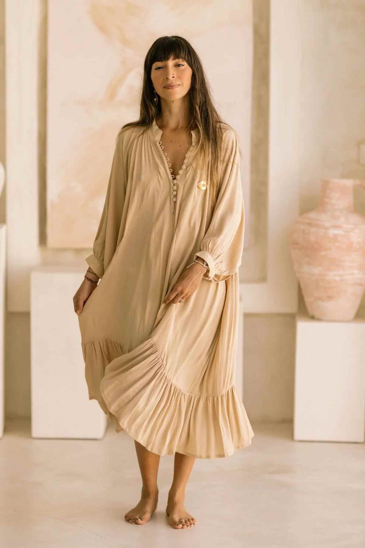 A woman stands barefoot in a softly lit room, wearing the flowing Bamboo Rayon FRIDA Gown Short by Myrah Penaloza, which features a pleated hem. The background showcases abstract art and pottery, capturing the serene elegance of handmade garments inspired by Frida.