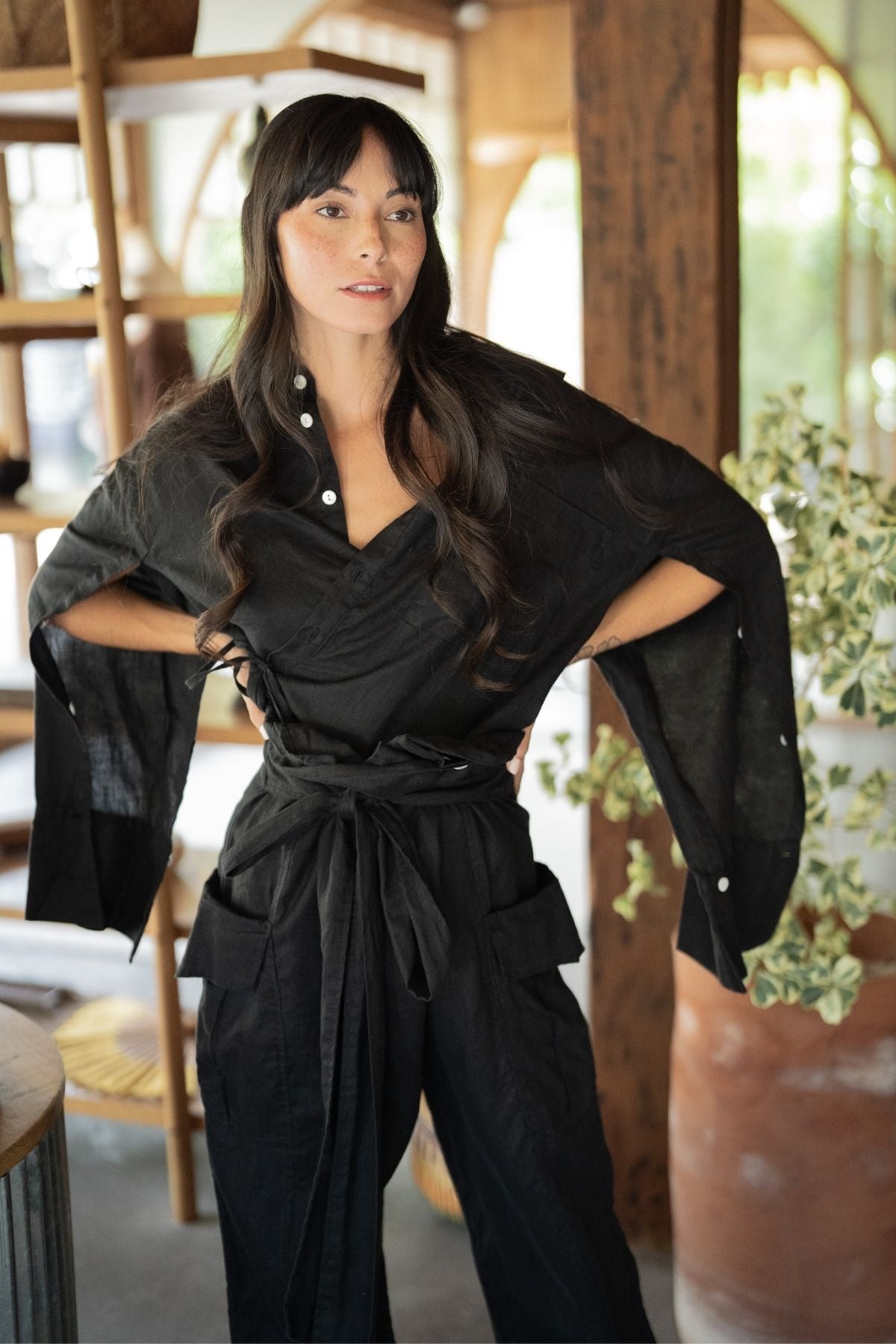 A woman with long dark hair and bangs stands confidently indoors, dressed in a stylish and sustainable black Sutra Linen Set (Made To Order) by Myrah Penaloza. The outfit features a belted waist and wide sleeves. She is looking slightly to the side with her hands on her hips. Behind her are wooden shelves, plants, and large windows.