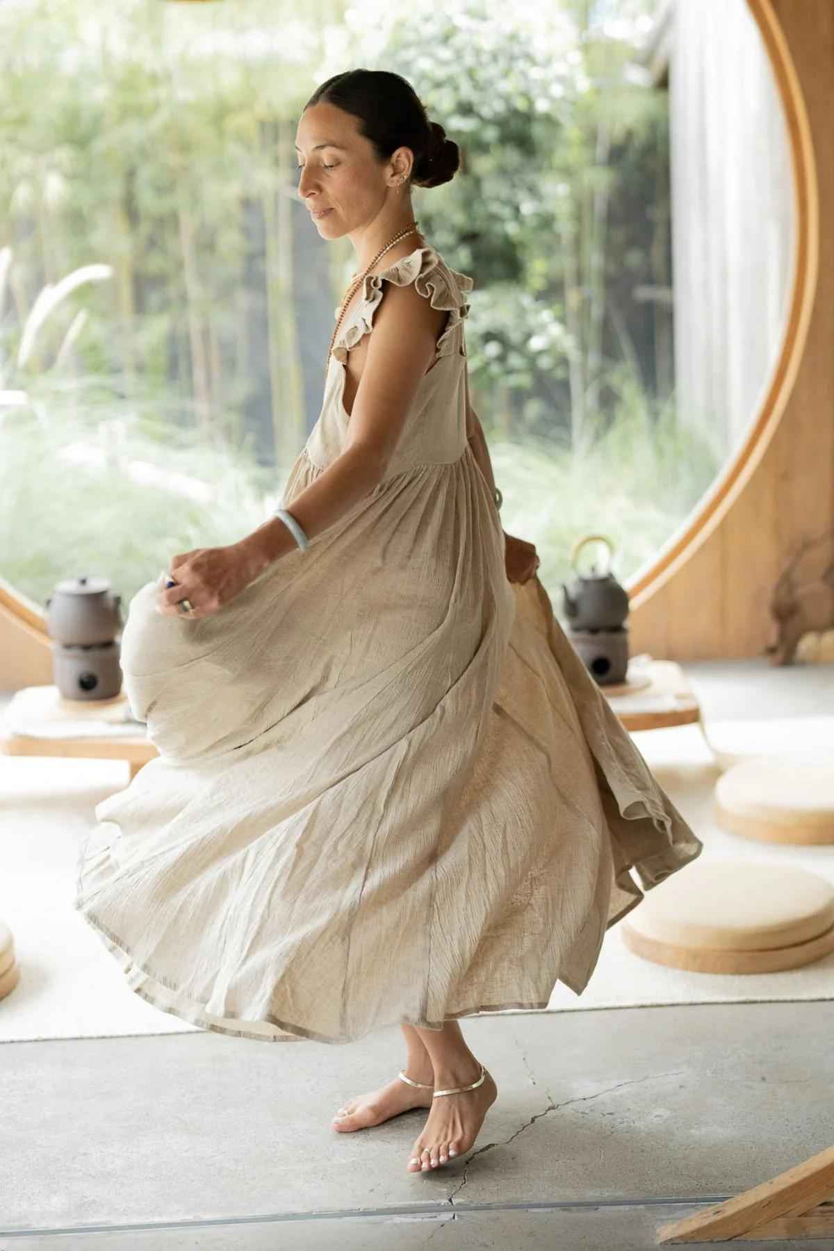 A woman in a flowing beige Linen Anna Yemaya Gown from Myrah Penaloza made of crinkle cotton dances barefoot on a concrete floor. She has her hair pulled back in a bun and is twirling gracefully. A round window with a view of greenery and bamboo provides a serene backdrop. Circular cushions are placed on the floor around her.