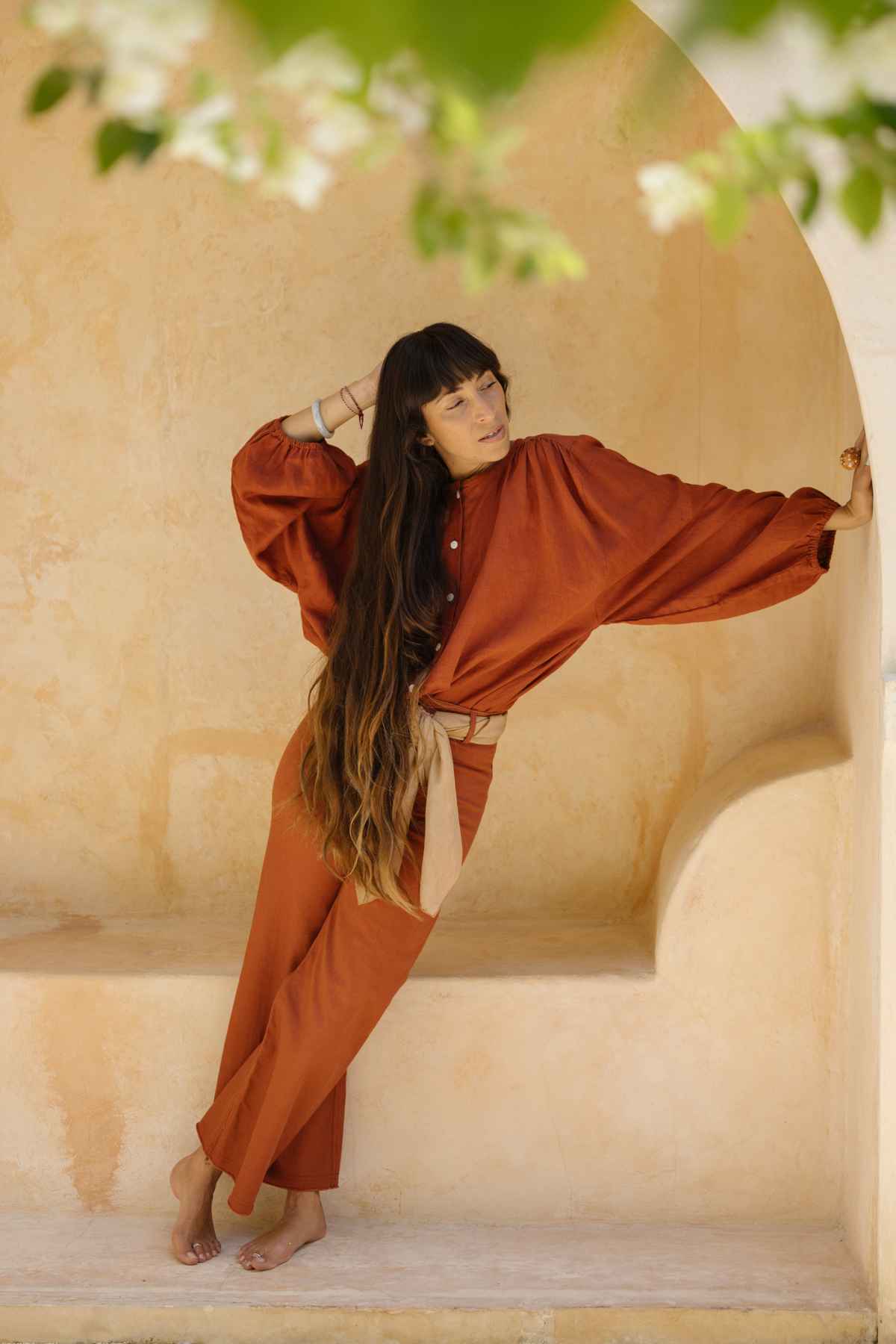 A woman with long, wavy hair leans against an archway in a beige, stuccoed space. She is wearing a flowy, rust-colored outfit with a belt at the waist and the Vida high waisted jeans by Myrah Penaloza, which are stonewashed and botanically hand dyed. Her expression is relaxed, and she is barefoot. The scene is serene and warm, with soft lighting.