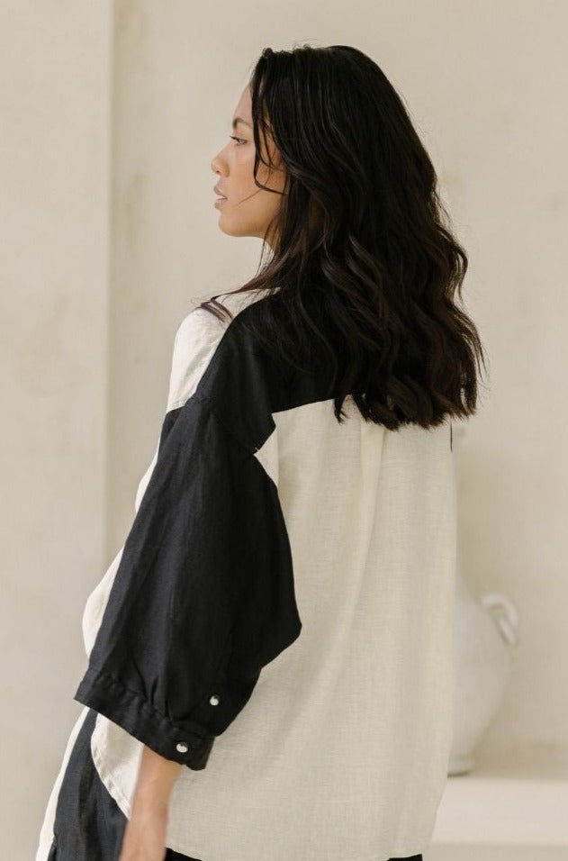 A woman with long, wavy dark hair is seen from the side and back, wearing a Myrah Penaloza Yin Yang Nidra Button Down Top that boasts a relaxed silhouette. The background is a plain, light-colored wall with a blurred white decor item visible in the corner.