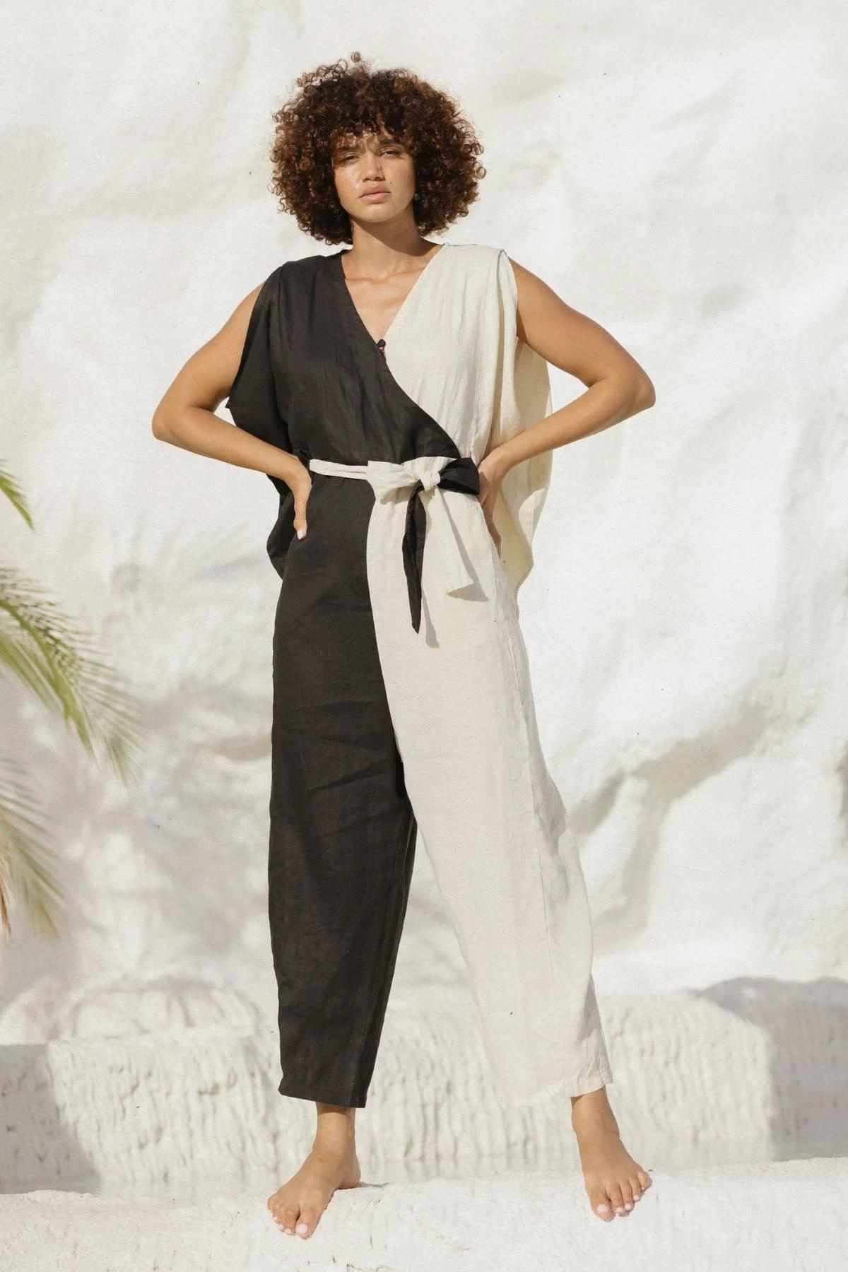 A person stands barefoot against a white background, wearing the French Linen Kuan Yin Playsuit by Myrah Penaloza. The sleeveless, two-toned outfit features a black left side and a white right side, cinched at the waist with an adjustable tie. One arm rests on their hip, and they have curly hair.