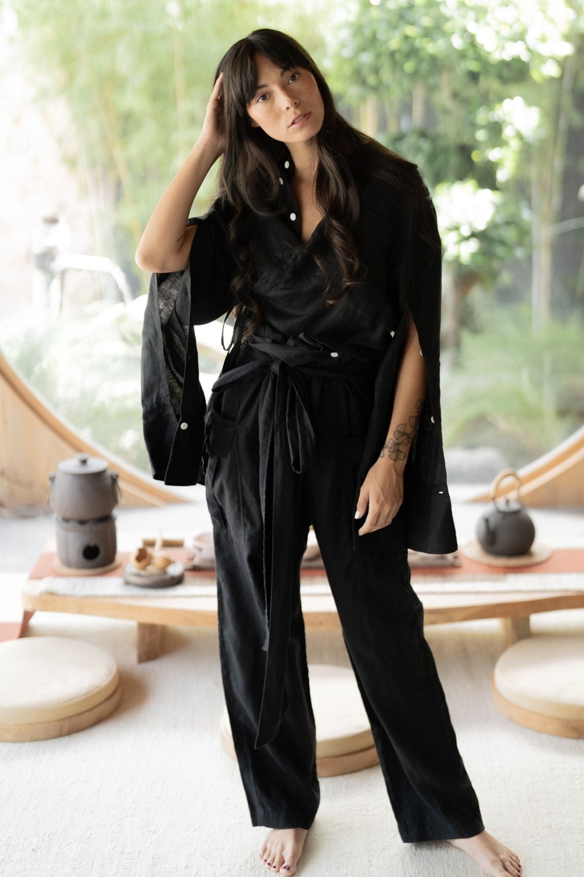 A woman with long dark hair, wearing the versatile black Sutra Linen Set by Myrah Penaloza, stands barefoot in a stylish, minimalistic room with circular windows. One hand rests on her head while the other hangs by her side. In the background, there is a low wooden table adorned with tea sets and surrounded by round cushions.