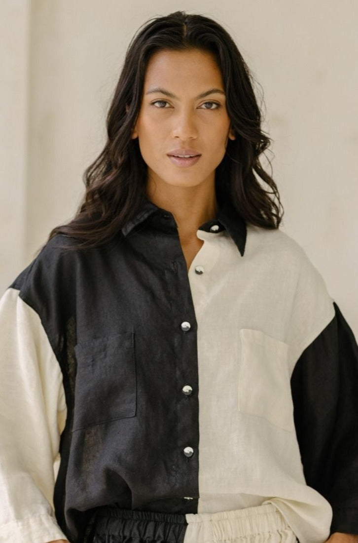 A person with long, wavy hair wearing a black and white Yin Yang Nidra Button Down Top by Myrah Penaloza is looking directly at the camera with a neutral expression. The background is plain and light-colored.
