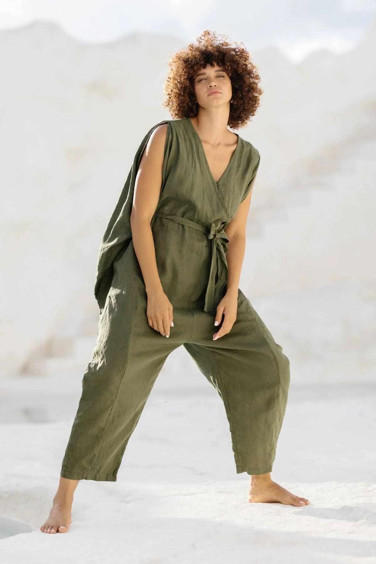 A person with curly hair stands barefoot on a white rocky surface, wearing the French Linen Kuan Yin Playsuit by Myrah Penaloza. The sleeveless, loose-fitting green linen playsuit features an adjustable waist tie. They exude a relaxed, confident posture amid light, natural rock formations in the background.