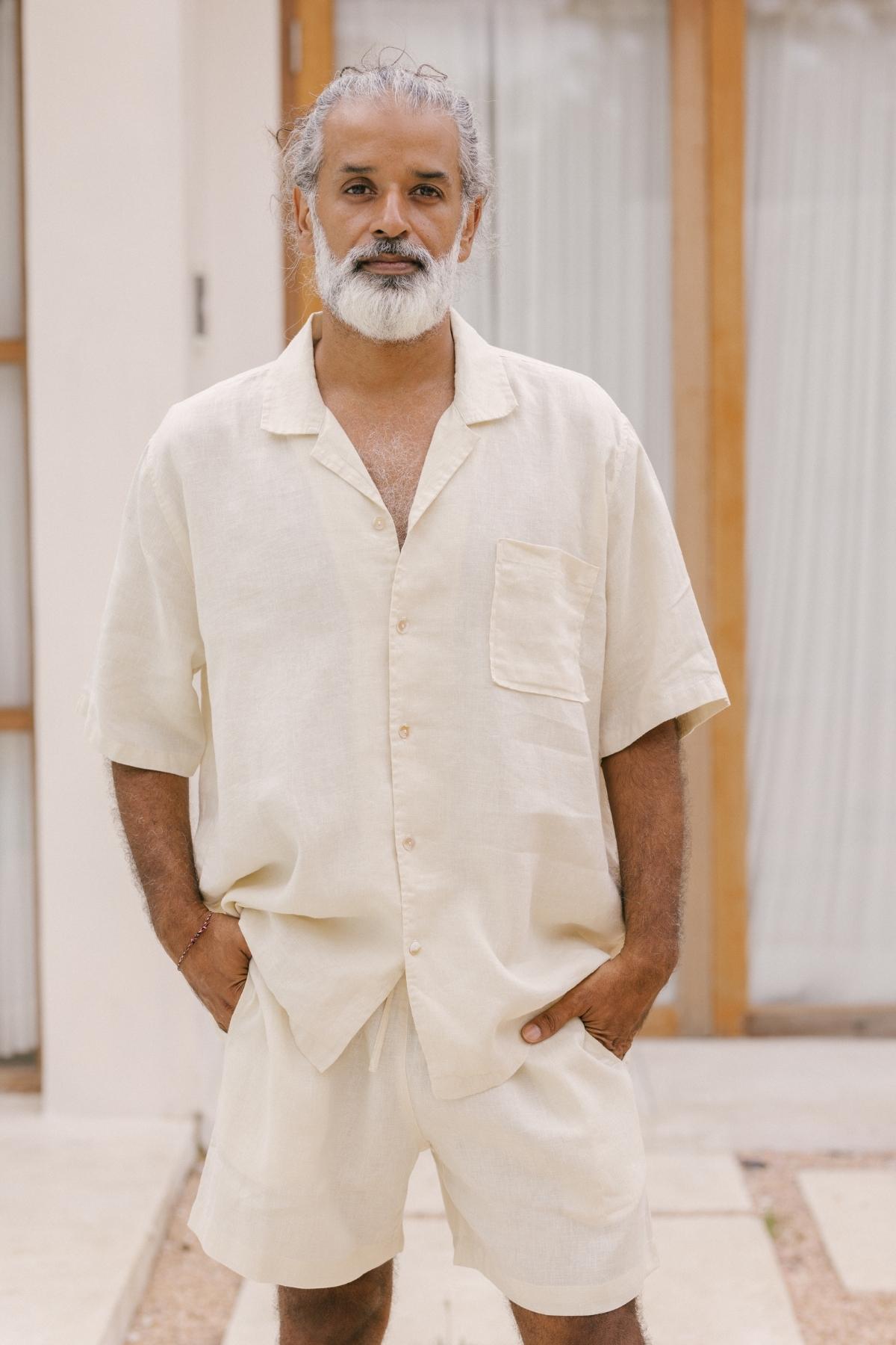 A man with gray hair and a beard stands outside wearing the lightweight and breathable Caravana Men's Linen Top & Short by Myrah Penaloza in beige. His hands are in his pockets as he stands in front of a building with large windows, capturing the relaxed essence of a nomadic traveler.