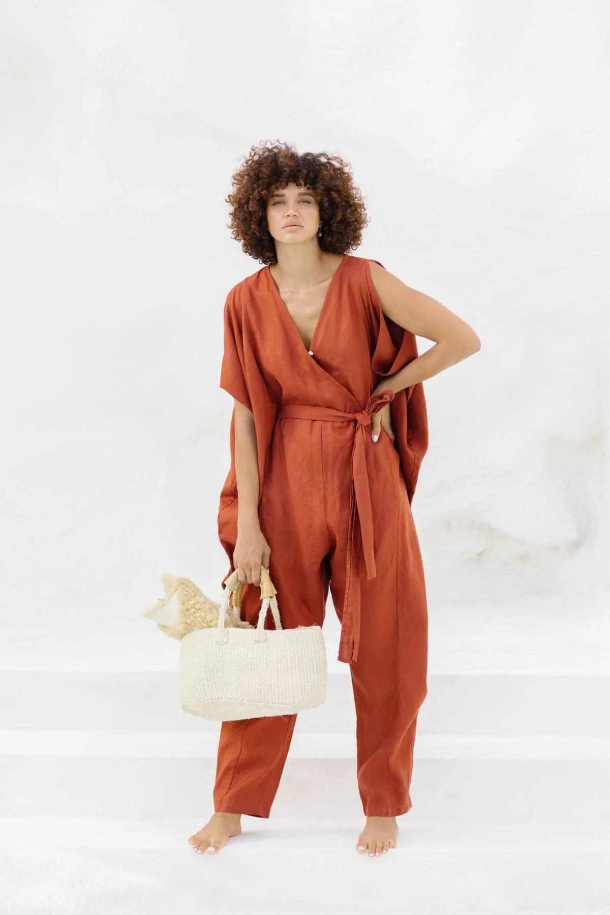 A person with curly hair stands barefoot against a plain background, wearing the orange French Linen Kuan Yin Playsuit by Myrah Penaloza, which features an adjustable waist tie. They hold a light-colored woven bag in their left hand.