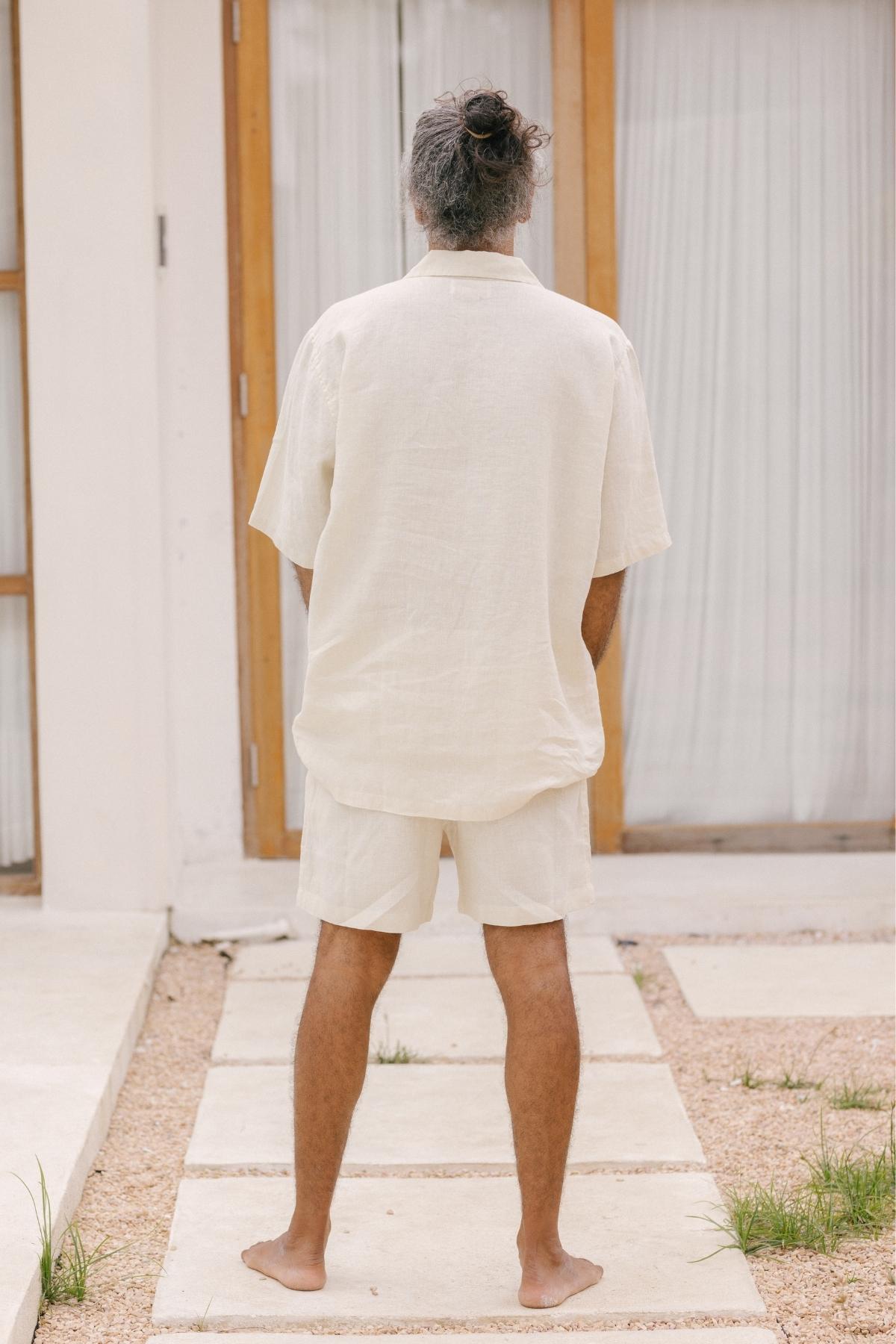 A nomadic traveler stands barefoot on stone tiles before a modern building with wooden-framed glass doors. Clad in a lightweight and breathable Caravana Men's Linen Top & Short by Myrah Penaloza in cream, their hair tied up in a bun, they face away from the camera, exuding effortless style.