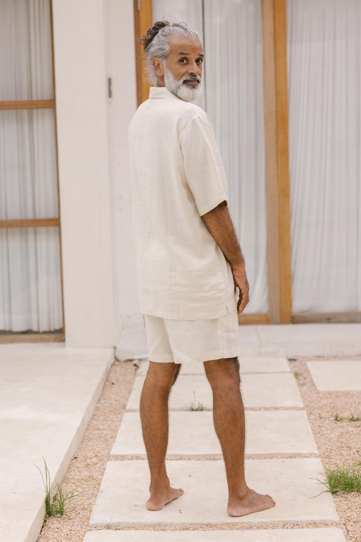 A man with gray hair and a beard stands barefoot on a stone path, wearing the lightweight and breathable Caravana Men's Linen Top & Short from Myrah Penaloza in beige. He gazes over his shoulder with a neutral expression. The background showcases white walls and glass doors adorned with curtains, capturing the essence of a nomadic traveler.
