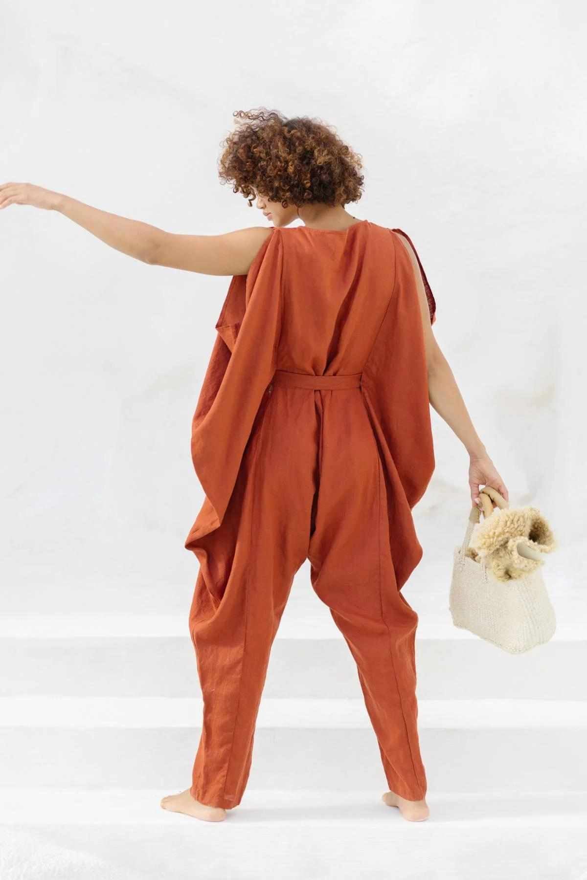 A person with curly hair is standing barefoot, facing away. They are wearing the French Linen Kuan Yin Playsuit by Myrah Penaloza, which features a loose fit and an adjustable waist tie in a rust color. In their left hand, they are holding a beige woven bag with fluffy contents. The background is a simple, light-colored wall.